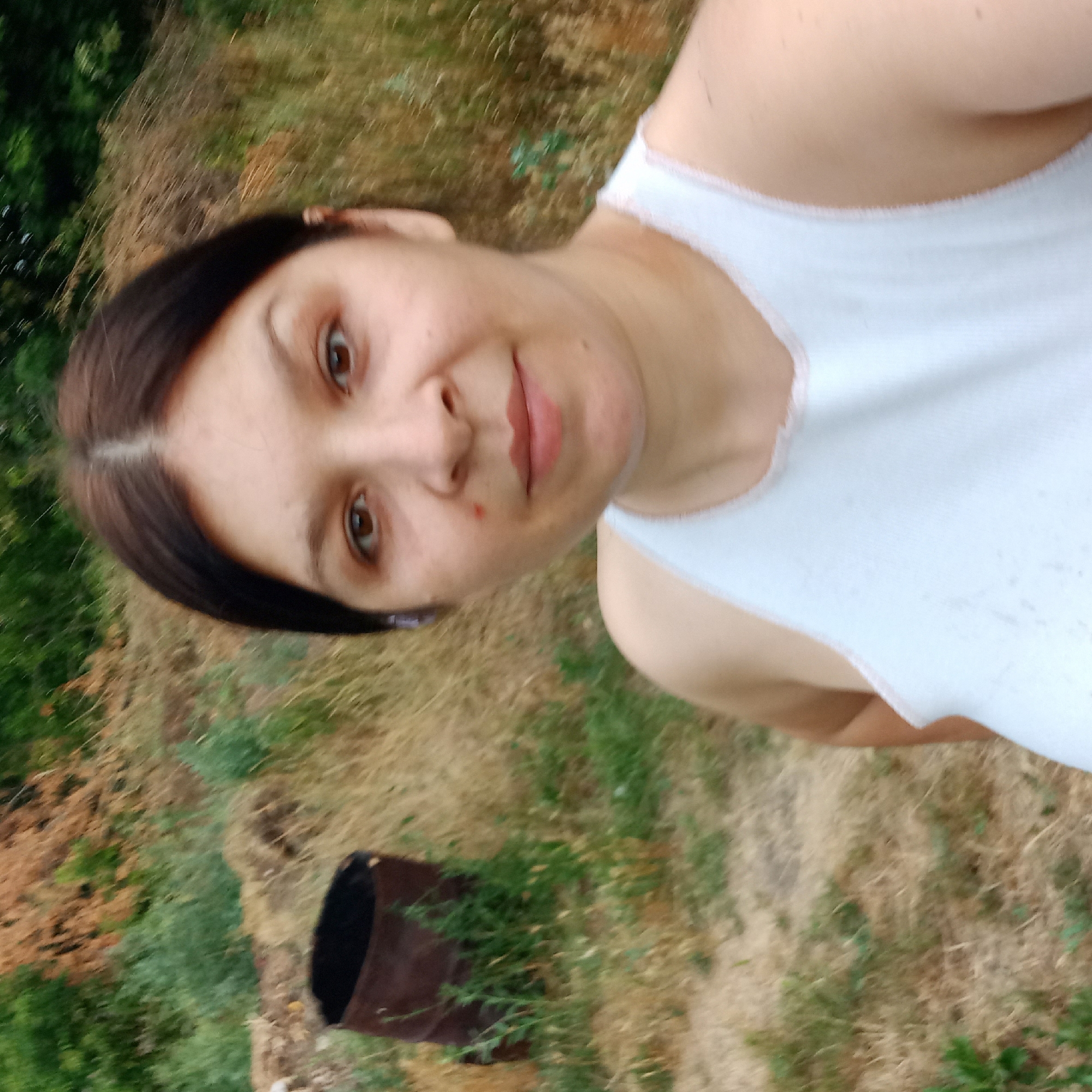 A selfie of a woman with brown hair and brown eyes is taken outdoors. She is wearing a white tank top. Behind her, a rusty barrel is visible on the left side of the image, and dry grass and green foliage fill the background. The scene appears to be in a rural or overgrown area, possibly within the city limits of Klyntsi, Ukraine.