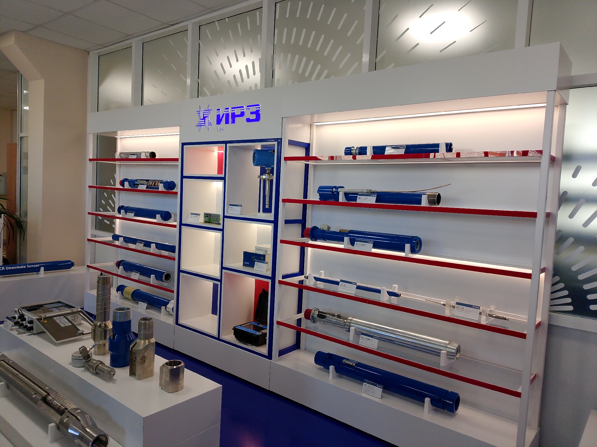 The image depicts a well-lit showroom or exhibition space displaying oil and gas industry equipment. The location is suggested by the "ИРЗ ТЭК" logo, which translates to IRZ TEC, a company based in Izhevsk, Russia.
The primary focus is on two white display units with integrated lighting. The unit on the left features open shelves presenting cylindrical blue and silver equipment. A separate section includes compartments showcasing smaller components and instruments. The right unit displays more cylindrical equipment of varying sizes on shelves with red accents. Some items are labelled with informational tags.
In the foreground, a white countertop showcases additional components, including metallic parts with threaded ends and other specialized connectors. The floor is a dark blue color.
In the background are tall windows with a frosted glass design, letting in natural light. A glimpse of foliage is visible through a doorway on the left. The overall environment appears to be a modern, professional showroom.