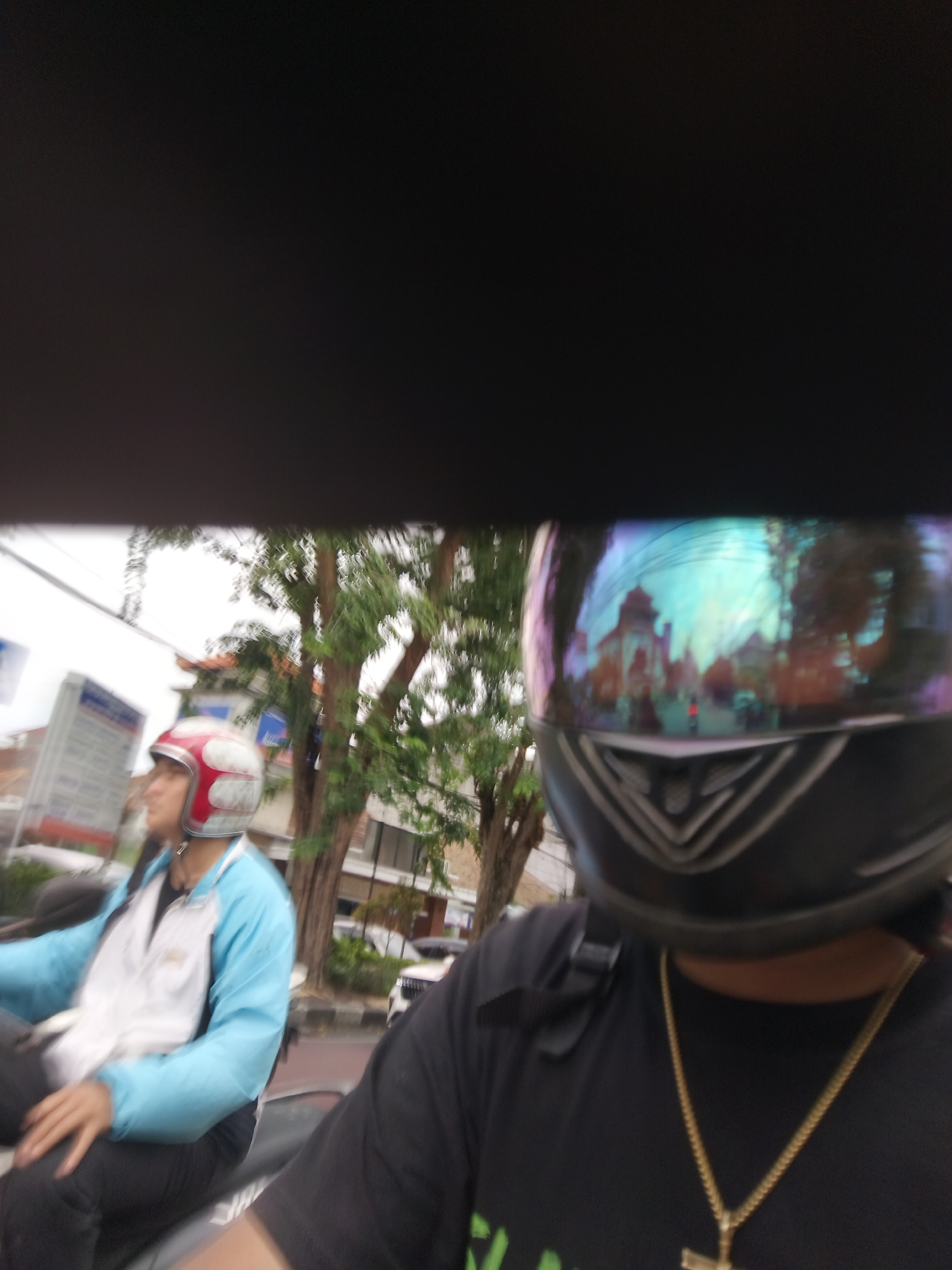 The image captures a close-up, first-person perspective of two individuals on a motorcycle in Denpasar, Indonesia. The rider in the foreground wears a black t-shirt and a helmet with a reflective visor displaying a colorful, distorted reflection of buildings and overhead wires. A gold chain with a cross pendant is visible on their neck. To the left, another person on the motorcycle wears a red and white helmet and a blue and white jacket, with their left arm resting on their lap. The background shows trees, buildings, and a street scene, suggesting an urban environment. The lighting suggests it is daytime, possibly overcast.
