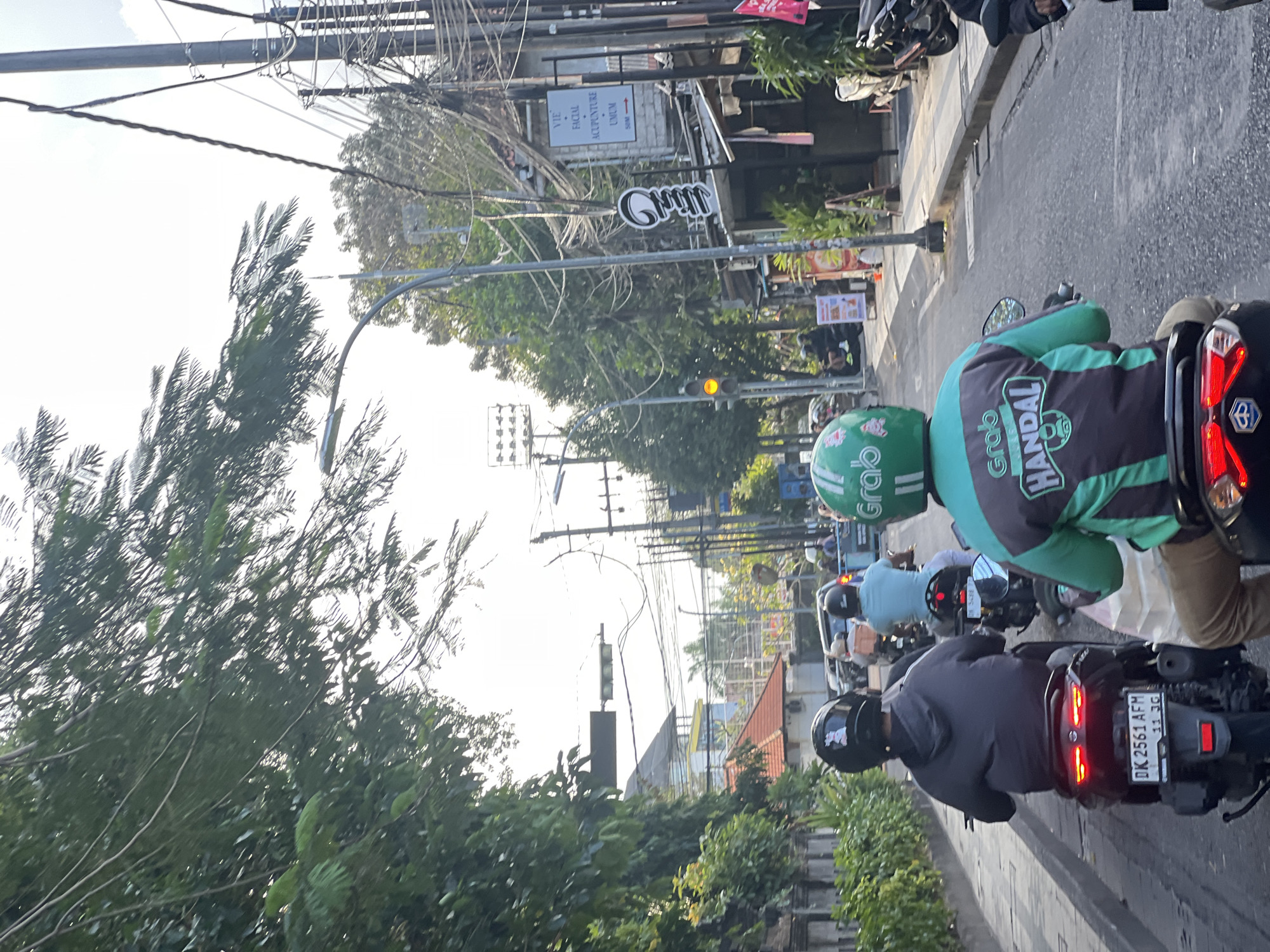 This is a street-level, eye-level, full-frame, vertical shot of a busy street in Kuta, Indonesia, with several motorbikes and riders. The perspective is from behind the riders, looking forward.
In the foreground, the backs of three motorbike riders are visible. The rider closest to the camera is wearing a dark grey hoodie and a black helmet with a white sticker of a rabbit on the back. Their motorbike is black and has a license plate that reads "DK 2561 AFM". To the right of this rider, another rider is wearing a green and grey jacket with "Grab" and "Handal" logos, and a green helmet. To the left and further ahead, a third rider is wearing a light blue shirt and a dark helmet.
The street is paved with asphalt and there are other vehicles and pedestrians further down the road. On the right side of the street, there is a building with a sign that says "VIE FACIAL ACUPUNCTURE UMUM". Above this, there are numerous electrical wires and poles, a common sight in Indonesian cities. On the left side of the street, there is a stone wall covered in green foliage, and further down, a building with a red-tiled roof. Trees with dense green foliage overhang the street from the left. The sky is bright and overcast. The overall impression is a typical urban street scene in Indonesia, bustling with activity and characterized by its unique infrastructure and tropical vegetation.