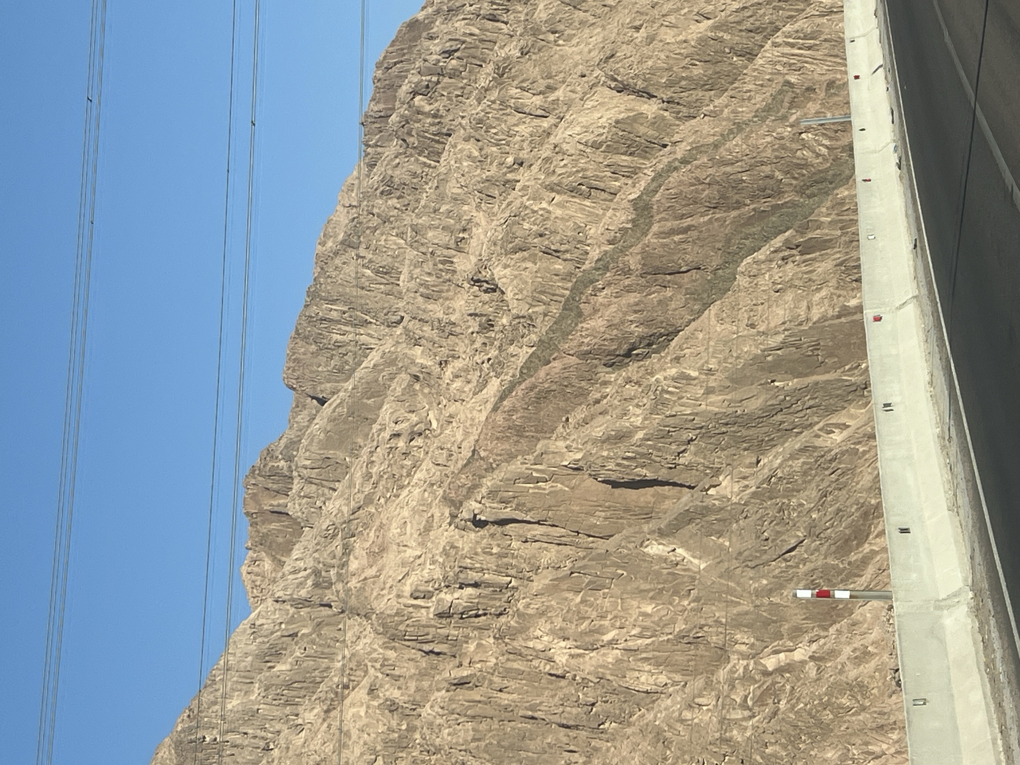 The image depicts a barren, mountainous landscape on a clear day, likely situated along a road in the vicinity of Dahab, Egypt, given the arid terrain and the presence of power lines common in such regions. The foreground features a paved road with a concrete barrier separating it from the rugged, rocky slope of a mountain. The mountain itself is characterized by steep, eroded faces displaying a rough, textured surface in various shades of brown and tan. Several power lines are visible stretching horizontally across the upper portion of the frame, with the sky being a vivid, cloudless blue. A red and white striped traffic marker is positioned on the side of the road, adjacent to the concrete barrier, and some debris is scattered along the base of the barrier. No people or animals are visible in the frame.