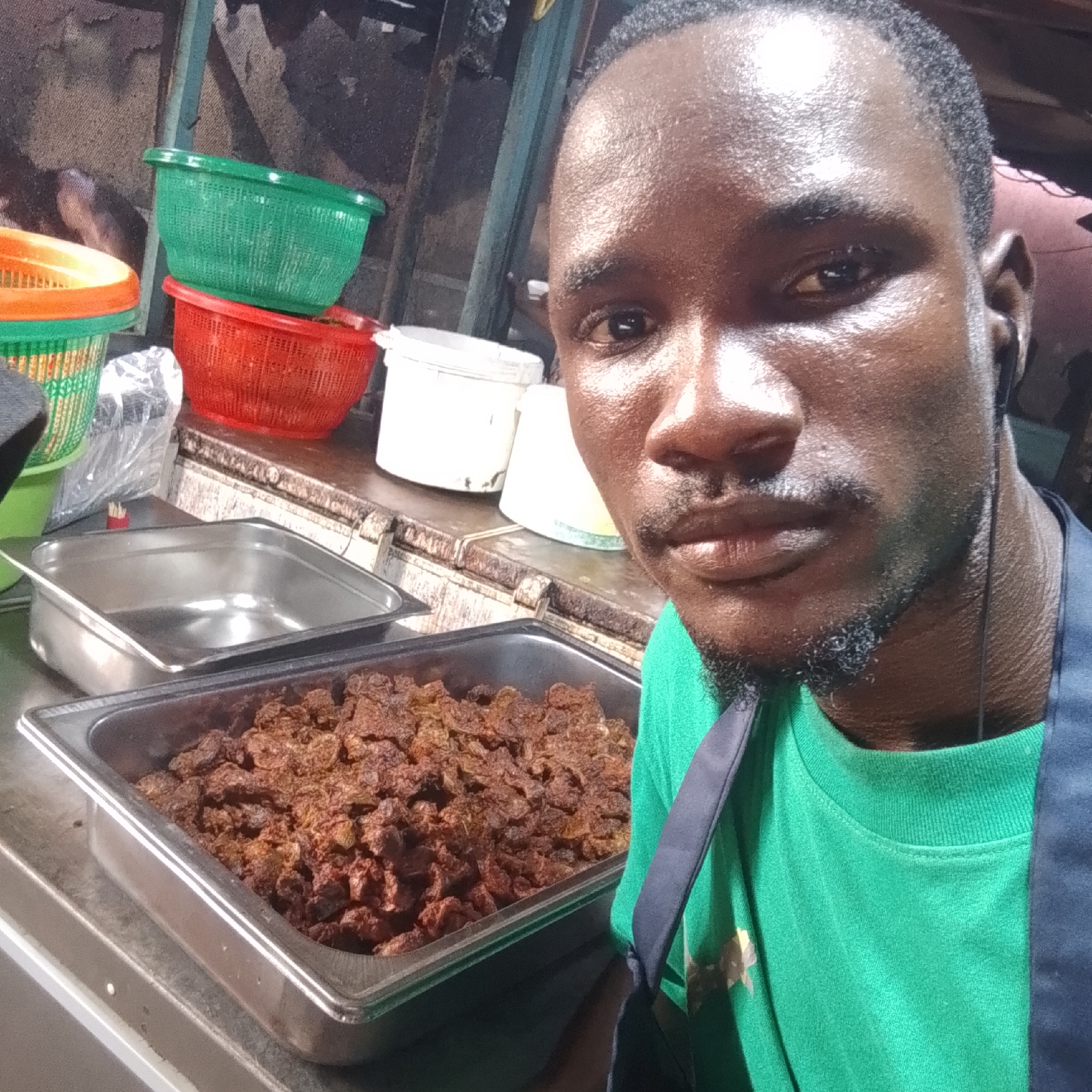 A close-up, medium shot captures a man from the shoulders up, positioned in what appears to be a food preparation or serving area in Lagos, Nigeria. The man, with dark skin and a light beard, is looking directly into the camera, his face glistening with sweat, suggesting a warm environment. He wears a bright green t-shirt. The background is cluttered and industrial, with various food service items visible. To the left, stacked plastic baskets in green and red are piled on a metal counter. A stainless steel food pan, containing small, brown, textured pieces of food, is in the foreground, directly in front of the man. Another dark-colored food pan is partially visible below it. The overall lighting is artificial and somewhat harsh, highlighting the textures of the man's skin and the food. The setting suggests a busy food stall or eatery.