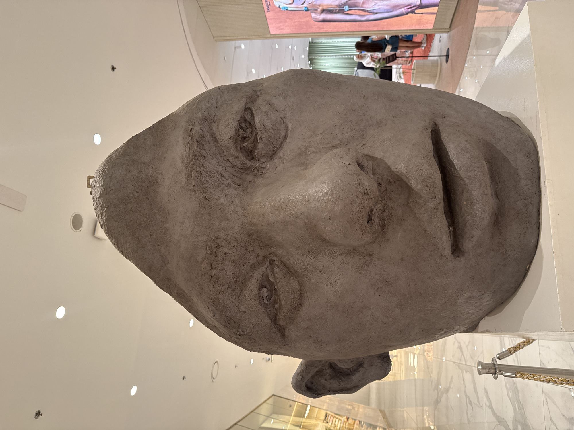A close-up, low-angle shot focuses on a large, abstract sculpture of a human head. The sculpture is made of a rough, textured gray material, resembling concrete or stone, with prominent features such as a large nose, slightly recessed eyes, and a simple mouth. The head is tilted upwards, and its left ear is clearly visible. The background is blurred, suggesting an indoor environment, possibly a public space or a gallery. The ceiling features recessed lighting. To the right, a vertical display screen shows a stylized human figure. In the middle ground, a few people are visible, appearing to be patrons or staff. A stanchion with a rope barrier is in the foreground to the left of the sculpture. The overall impression is that of an art installation within a modern, well-lit commercial or exhibition space.