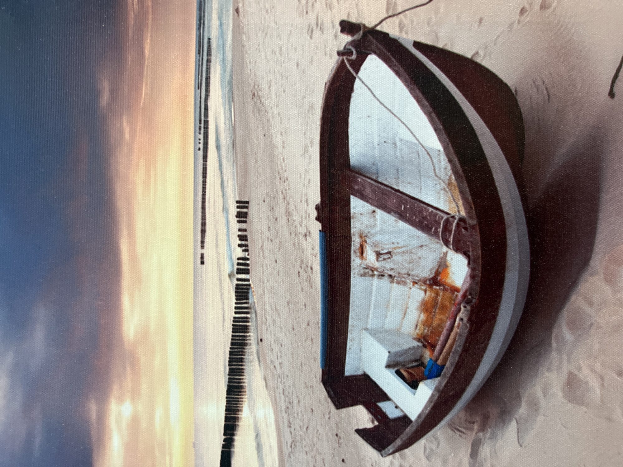 A small, weathered wooden rowboat rests on a sandy beach. The boat is painted dark red on the outside with a white stripe running along the gunwale and a white interior. The interior shows signs of wear and tear, with some rust stains and a wooden crossbeam. A blue object and some wooden poles are visible inside the boat. A rope extends from the bow of the boat to the right. The beach is made of light-colored sand with footprints visible. In the background, the sea meets the sky at the horizon. A series of wooden posts, likely breakwaters, are planted in the shallow water along the coast. The sky is dramatic, with dark clouds in the upper portion transitioning to lighter, pastel colors of pink, orange, and yellow near the horizon, suggesting either sunrise or sunset. There are no people visible in the image. The scene depicts a serene, coastal environment. The image does not contain any information that allows for its location in the city of Eretria Municipal Unit, Greece.