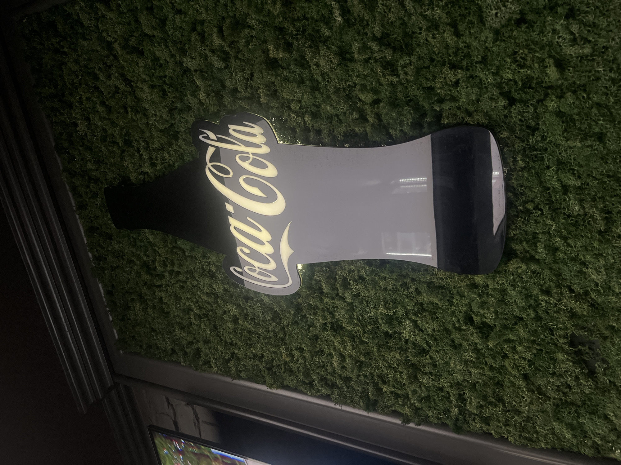 An illuminated Coca-Cola bottle-shaped sign is centrally positioned against a textured wall covered in dense, dark green, moss-like material. The "Coca-Cola" script, rendered in white, glows with a warm yellow light, creating a halo effect around the letters against the dark grey silhouette of the bottle. Reflections of what appear to be vertical LED light strips are discernible on the lower, darker portion of the sign. To the upper left, a dark, ribbed architectural molding runs horizontally, connected to a vertical dark frame. The overall lighting is dim, emphasizing the illuminated sign against the dark, textured background. The scene depicts an interior or semi-enclosed commercial space. The specific location in Burgas, Bulgaria, cannot be ascertained from the provided visual data.