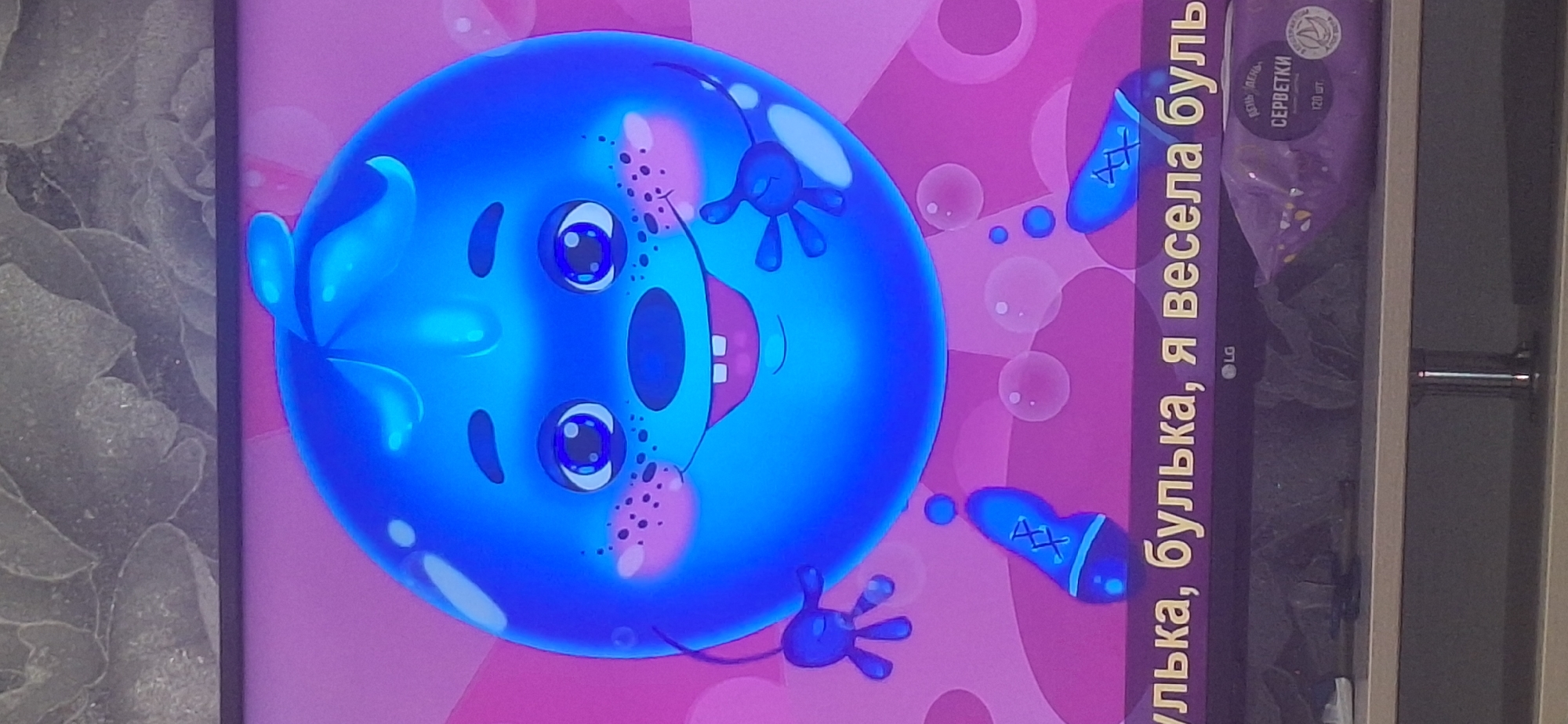 The image shows a cartoon character that appears to be a blue, anthropomorphic bubble with a face, arms, and legs. The character is depicted against a pink and purple background with some bubbles. Below the character, there is text in yellow that appears to be Russian: "Булька, булька, я весела буль". There is also an LG logo on the screen. Below the screen, a package of napkins with Russian text ("СЕРВЕТКИ") and "120 шт." is visible. The image is likely a screenshot or photograph taken from a screen. There is no indication of the location being in Bila Tserkva, Ukraine.