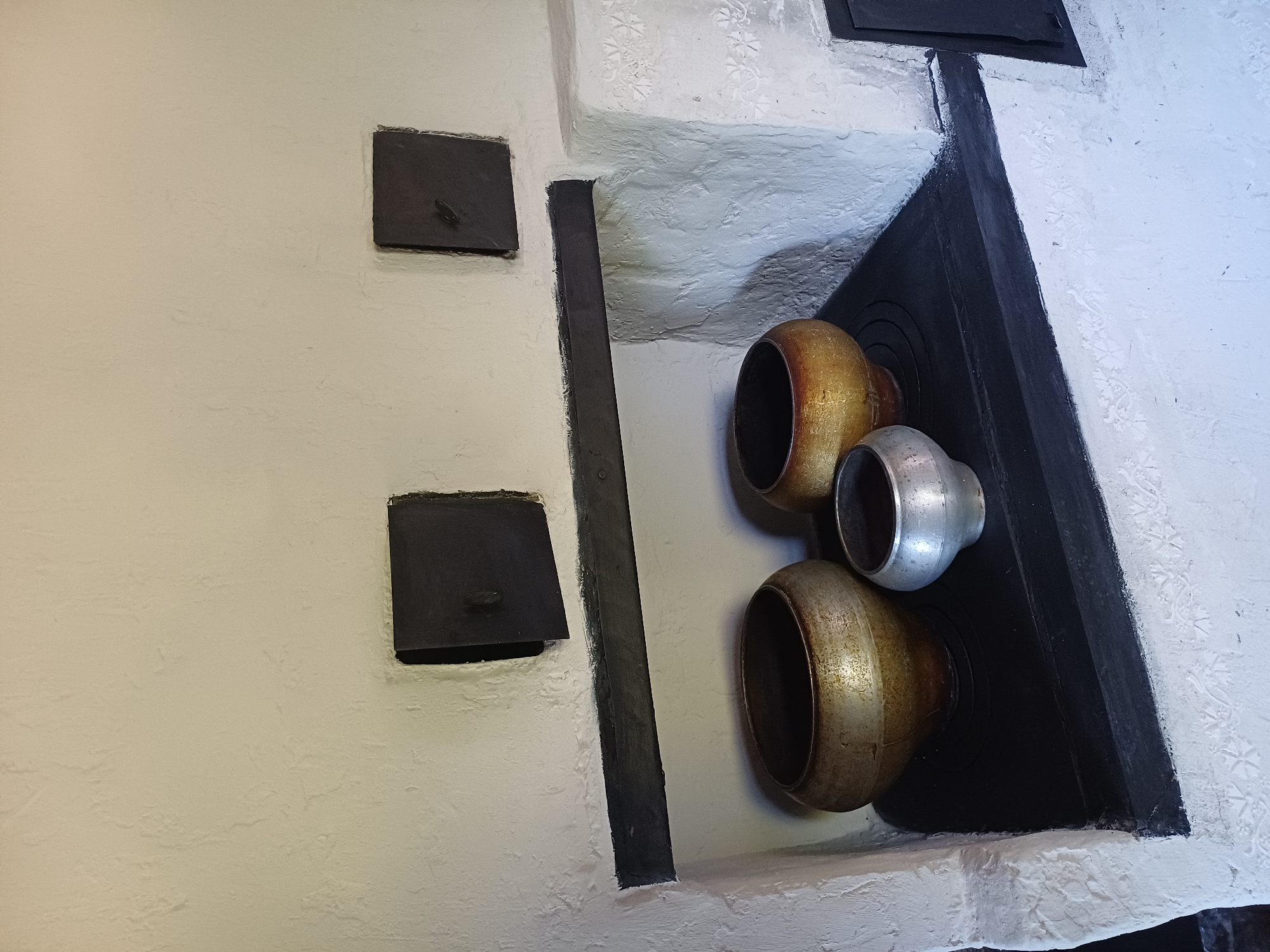 The image displays an interior view of a rustic, white-plastered stove with three vintage pots placed on its top surface. The stove has black metal accents, including two square openings with handles above a horizontal shelf, and a darker rectangular area on the right side. The pots vary in size and color, with two larger, aged metal pots and a smaller, silvery one in the front. The stove and its surroundings exhibit a textured, rough plaster finish, suggesting an older construction. The scene is lit from above and to the right, casting subtle shadows. This image was captured in Sulymivka, Ukraine, likely within a historic or traditional dwelling.