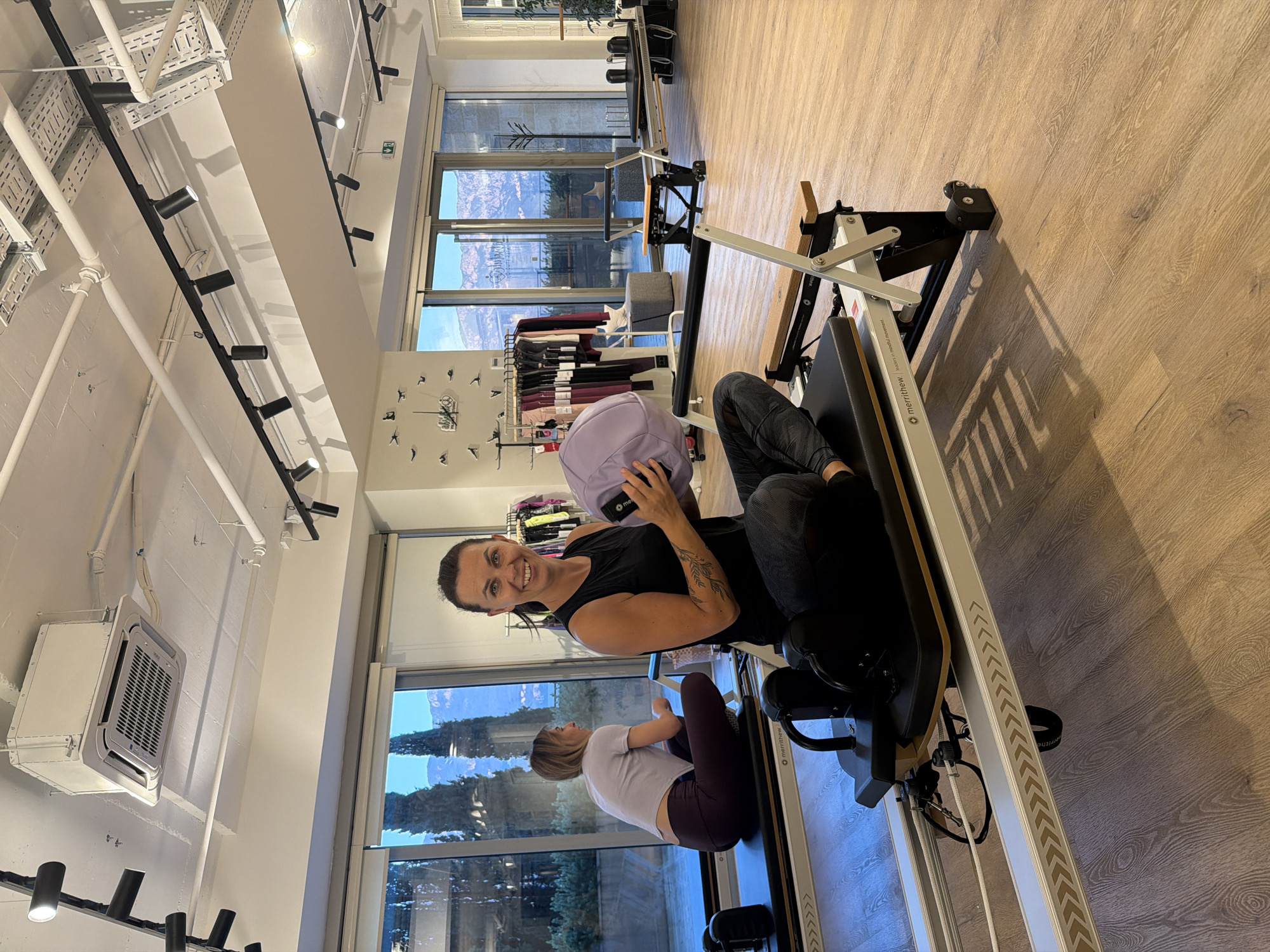 In a Pilates studio in Boreti, Montenegro, a woman with dark hair pulled back smiles at the camera while holding a light purple ball. She is seated on a Pilates reformer, wearing a black sleeveless top and patterned leggings. Another woman in a grey top and dark leggings is seated on a similar reformer in the background, facing away from the camera. The studio has large windows overlooking a scenic landscape with mountains and trees. The floor is light wood, and the ceiling features track lighting and an air conditioning unit. Racks of athletic clothing are visible in the background, as are other Pilates reformers.