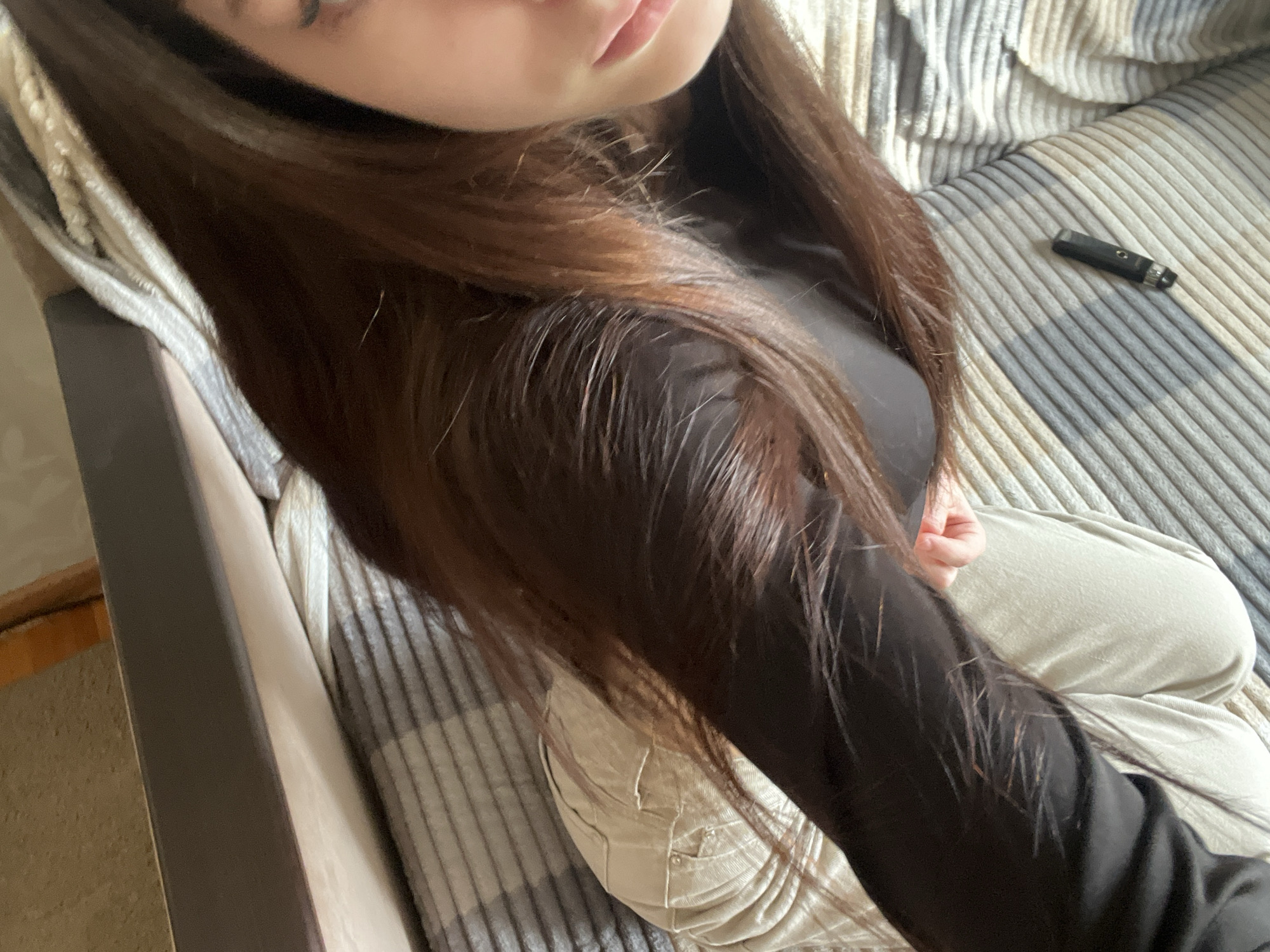 The image depicts a person seated on a textured, striped surface, possibly a sofa or chair. The person is wearing a long-sleeved black shirt and light-colored pants. A black, rectangular object, possibly a vape pen or similar device, is visible on the striped surface. The person has long, dark brown hair. The location is unspecified in the city of Unknown, Russia. No specific actions or interactions are evident. The environment is indoors.