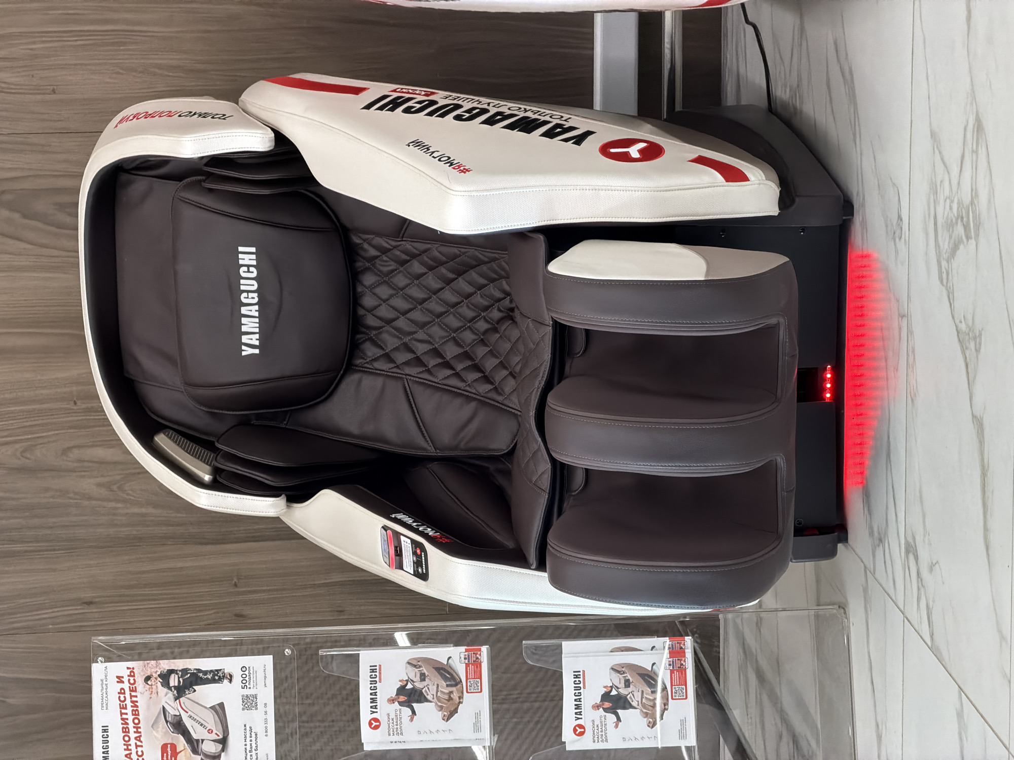 The image captures a premium YAMAGUCHI massage chair displayed in what appears to be a showroom or retail space in Izhevsk, Russia. The chair, predominantly white and dark brown/black, features a sleek, modern design with plush, leather-like upholstery, including a quilted pattern on the backrest. Red accent stripes and the YAMAGUCHI logo are prominently displayed on the chair's headrest and side panels.
The chair is situated on a light wooden floor, adjacent to a wall with a white and gray marble-like pattern. Red LED lights emit from the base of the chair, illuminating the area behind it and adding to its contemporary aesthetic.
In the foreground, a clear acrylic display stand holds several brochures advertising YAMAGUCHI massage chairs. The flyers show images of people relaxing in the chairs and feature various promotional texts. Visible text includes the brand name "YAMAGUCHI" repeated multiple times, along with Russian phrases such as "ТОЛЬКО ЛУЧШЕЕ" (Only the best), "ПРЕМИАЛЬНЫЕ МАССАЖНЫЕ КРЕСЛА" (Premium massage chairs), "ОСТАНОВИТЕСЬ И УСТАНОВИТЕСЬ!" (Stop and settle in!), and "Японский МАССАЖ ДЛЯ ВАШЕГО ДОЛГОЛЕТИЯ" (Japanese massage for your longevity). The hashtag "#ЯМОГУЧИЙ" is also visible. One brochure mentions "5000 в подарок" (5000 as a gift), a phone number (8 800 333-56-08), and the website yamaguchi.ru. Japanese text "ロングライフ" (Long life) is also present. The scene suggests a well-lit indoor environment, likely during business hours, showcasing the product to potential customers. There are no people visible in the image.