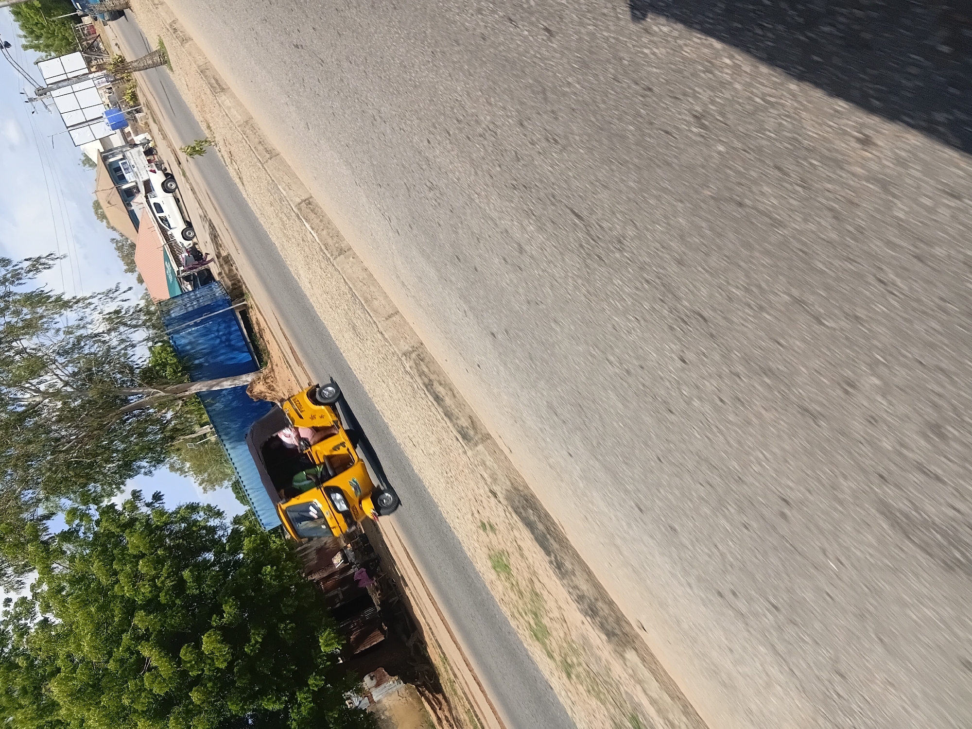 A yellow motorized tricycle, known locally as a "keke napep," is traveling on an asphalt road in Jalingo, Nigeria. A person wearing a pink garment is visible in the passenger seat of the tricycle. The road is bordered by a sandy verge and a concrete curb. In the background, there are several buildings, a large blue shipping container, and lush green trees. The sky is partly cloudy. The image appears to have been taken from a moving vehicle.