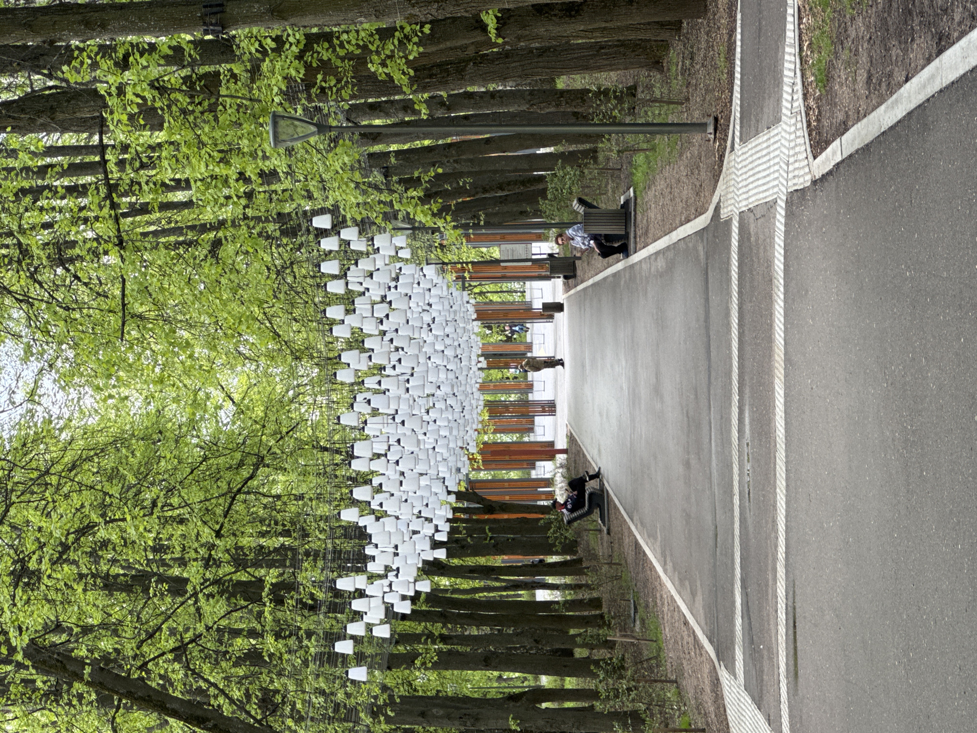 The image shows a park or pedestrian zone in Nizhny Novgorod, Russia. A straight paved path extends into the distance. Along one side of the path is a line of trees with young green leaves, providing shade. Benches are placed intermittently along the path, and some people are sitting on them. Suspended above the path between the trees are numerous white, lamp-shaped objects arranged in a grid-like pattern. A lamppost is visible near one of the benches. The weather appears to be overcast.