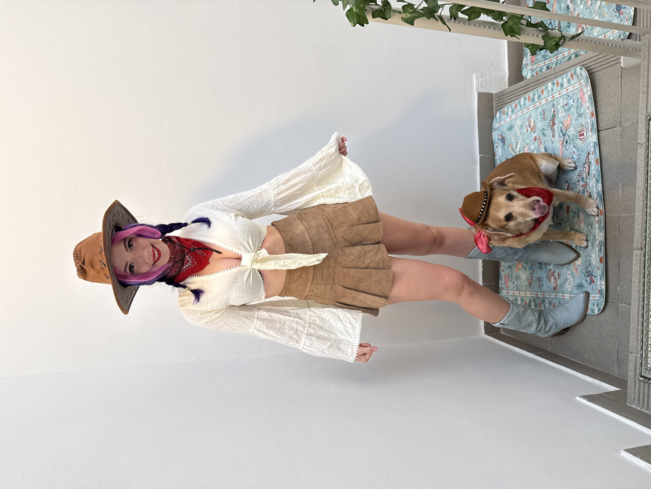 A woman and a dog pose indoors dressed in cowboy attire. The woman wears a tan cowboy hat, a white blouse tied at the waist, a tan skirt, and light blue cowboy boots. The dog is wearing a small cowboy hat and a red bandana. They both appear to be standing on a white floor in front of a staircase with patterned mats on the steps. Green plants are visible at the top of the staircase.