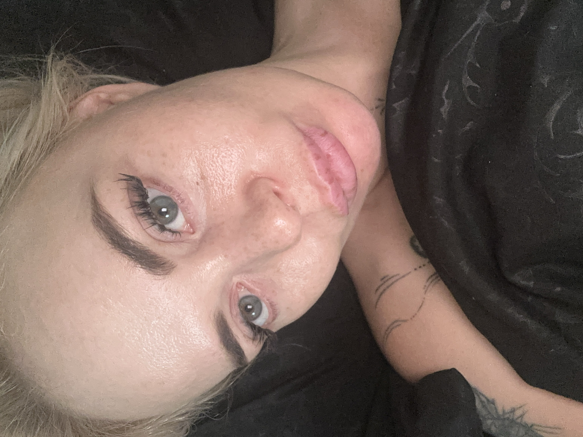 A close-up, overhead shot shows a fair-skinned person with blonde hair, lying down and looking at the camera. The person has light blue eyes, dark eyebrows, and long eyelashes. There are freckles on their cheeks and nose. Their lips are slightly parted, revealing a small bruise on the upper lip. They are wearing a black shirt or blanket, which is wrinkled and covers their chest and arm. There are tattoos visible on their arm and shoulder. The background is dark and out of focus. The image appears to have been taken indoors, possibly in a bedroom. The location is unknown.
