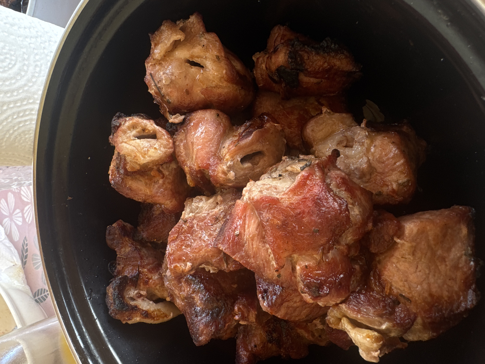 A close-up, eye-level shot shows a black ceramic pot containing a heap of roasted meat pieces. The meat appears to be pork, characterized by its reddish-brown, slightly charred exterior and varied shapes. Some pieces have a hollow interior, suggesting they may have been skewered during cooking. The pot is positioned on a surface with a hint of other objects visible on the left, including what appears to be white paper towels and a patterned fabric. The image's location is unknown, and there is no evidence to suggest the photograph was taken in the city of Chaikovsky, Russia. The image focuses exclusively on the cooked meat within the pot, with no people, actions, interactions, or specific environmental details depicted.