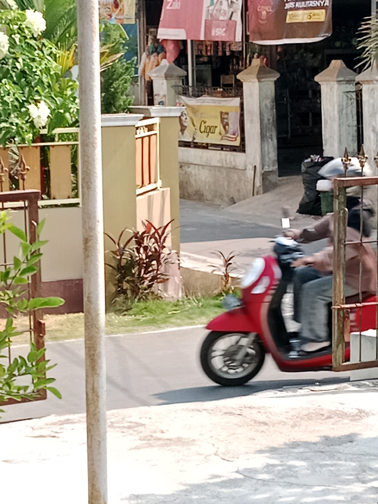 In Waru, Indonesia, a person is riding a red scooter. The scooter is in motion on a paved road, and the rider is wearing jeans and a light brown jacket. Behind the scooter, a large black garbage bag is visible. The scene is outdoors, with a building and foliage in the background. A white pole stands in the foreground, partially obscuring the view. A metal fence with decorative elements is also present on the right side of the image.