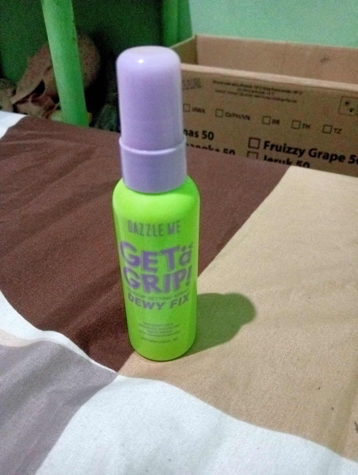 A neon green spray bottle with a light purple cap stands upright on a brown and beige striped surface, possibly a bedspread or blanket. The bottle has text printed on it in pink and white. The visible text on the bottle reads "DAZZLE ME", "GET GRIP!", and "DEWY FIX". The bottle is casting a distinct shadow to its right. In the background, a portion of a cardboard box with text and checkboxes is visible. The overall scene appears to be indoors. There is no visible indication of the city of Kota Negara Timur, Indonesia.