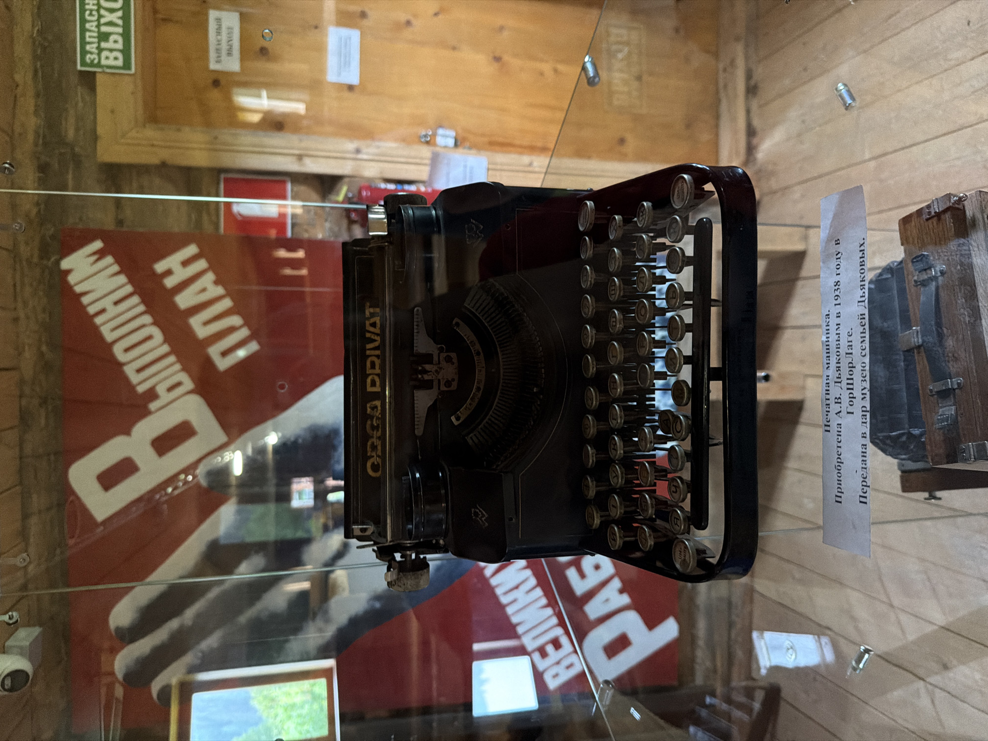 A vintage black typewriter is displayed on a glass shelf in a museum. The typewriter has a metal frame, black keys with Cyrillic lettering, and a carriage return lever. Behind the typewriter, a red banner with white lettering and a large gray hand graphic is visible. The text on the banner appears to be in Russian. To the right of the typewriter, a wooden door with a sign reading "ЗАПАСН. ВЫХОД" (Emergency Exit) is visible. The floor beneath the display is made of light-colored wood planks. A small descriptive plaque is placed in front of the typewriter, also on the glass shelf. The plaque contains Russian text. The overall environment suggests an indoor exhibition space with rustic wooden walls.