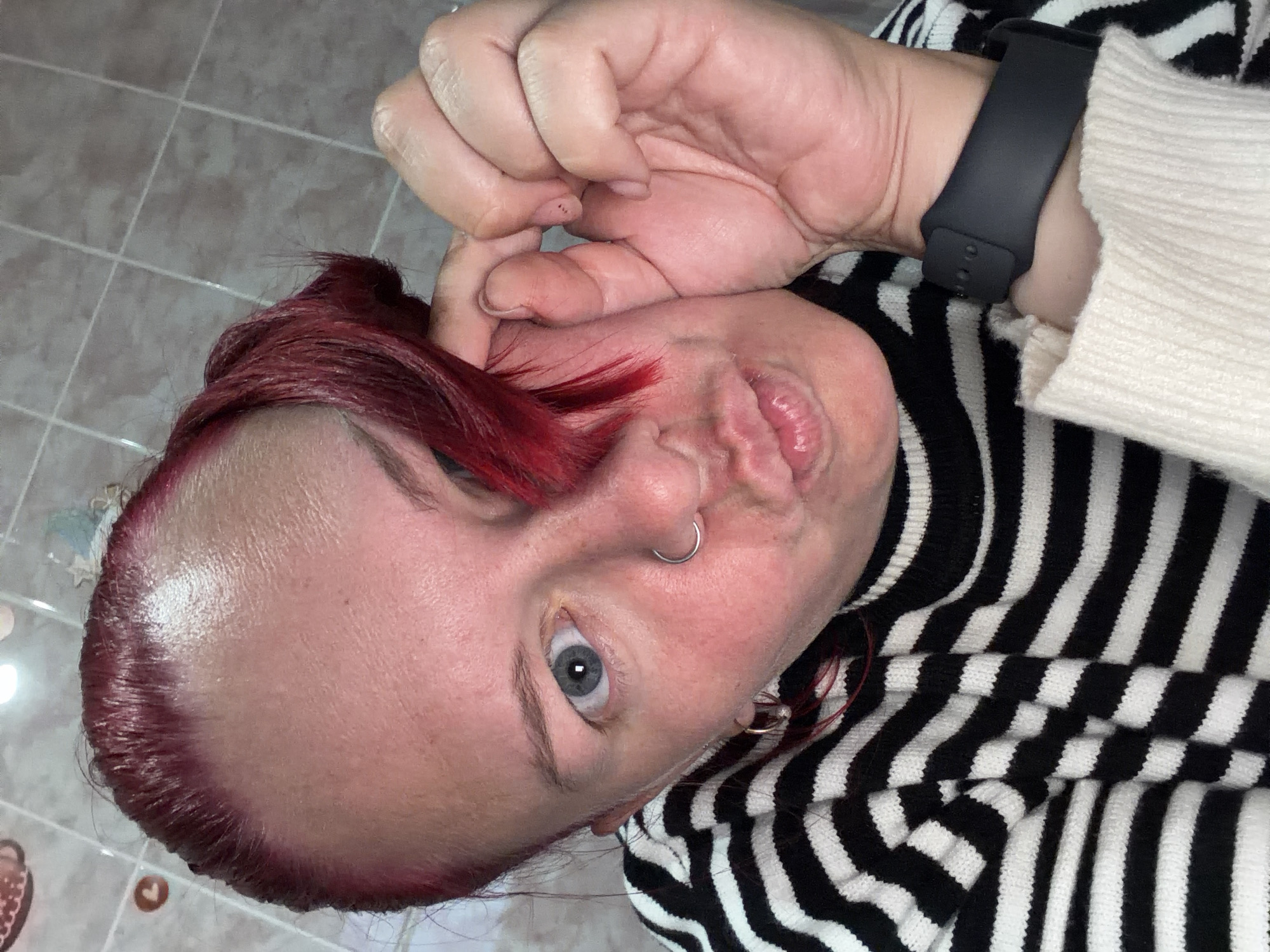 A close-up selfie shows an individual with bright red-dyed hair, fair skin, and light blue eyes. The person has a silver hoop nose ring in their left nostril and is wearing a black and white striped knitted top. On their left wrist, a black digital fitness tracker or smartwatch is visible. Their lips are puckered in a playful or exaggerated expression, and their left eye is wide open, looking directly into the camera. A hand, possibly the person's own, is partially visible near the top of their head, appearing to hold back hair or rest on the temple. The background consists of a tiled floor with light grey or beige square tiles and darker grout lines. The image is taken indoors, with artificial lighting casting a slight sheen on the person's forehead. No text is visible.