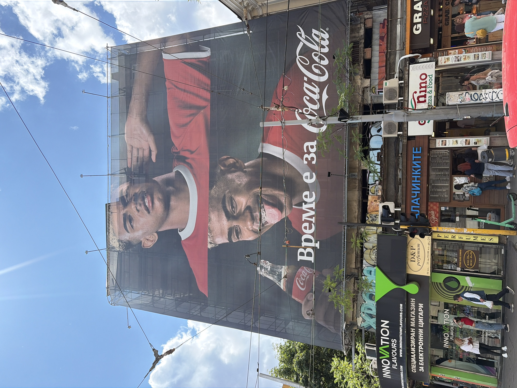 The image shows a large billboard for Coca-Cola featuring two close-ups of a young man. The billboard is positioned above a street in Sofia, Bulgaria. In the lower portion of the image, several storefronts are visible, including "INNOVATION FLAVOURS," "D&P perfumium," "NINO Pizza & Food," and "GRAB PIZZA." People are walking on the sidewalk in front of the stores. Overhead, electrical wires crisscross the scene, and trees grow along the street. The sky is blue with scattered clouds.