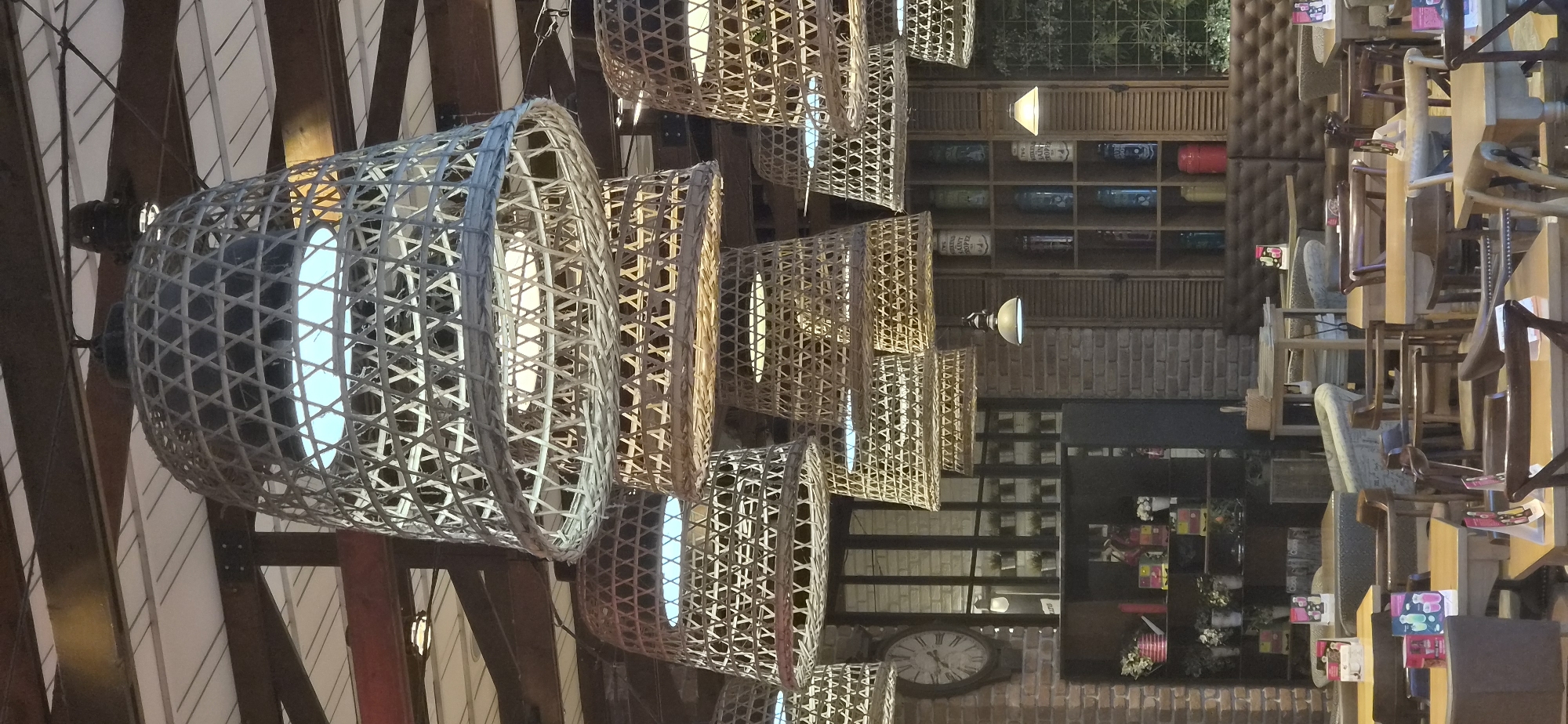 The image shows an indoor scene, likely a restaurant or cafe, in Varna, Bulgaria, recognizable by the architectural style and interior design elements. The focal point is a series of wicker basket-style light fixtures suspended from the ceiling. These fixtures are arranged in a row, varying in size and shade, with glowing bulbs visible inside. The ceiling structure appears to be exposed, with wooden beams and a white-paneled backdrop.
In the background, a wooden shelving unit displays various items, including bottles and decorative objects. To the right, there are tables and chairs, some stacked or arranged close together, suggesting the area may be prepared for service or in a state of transition. The seating area seems to incorporate a mix of seating styles, including cushioned chairs and wooden chairs.
A large clock is visible on the brick wall in the background. The overall ambiance suggests a blend of rustic and modern design elements, creating a warm and inviting atmosphere.