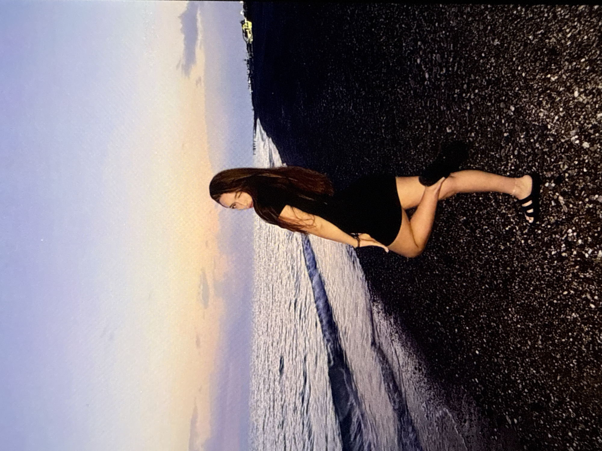 A young woman with long, dark hair is posed on a pebble beach at dusk. She is wearing a short, black dress and black sandals. The ocean is to her left, with gentle waves lapping the shore. The sky above is a gradient of purple and orange, indicating the setting sun. In the background, along the coastline, are a few buildings with lights on, suggesting a settlement. The overall scene captures a moment of peaceful solitude by the sea. The location is not specified in the image.