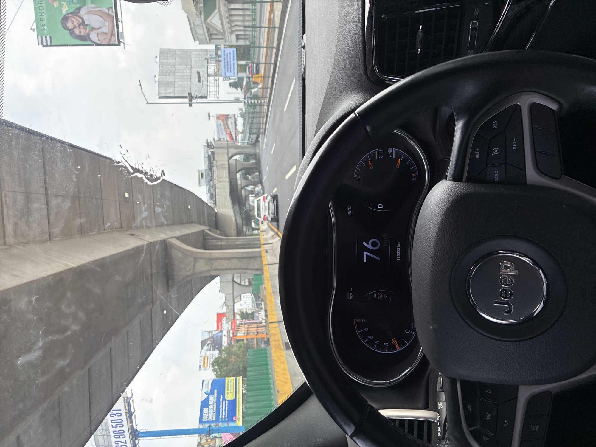 The image shows the view from inside a moving Jeep vehicle, likely on an elevated roadway or bridge in Naucalpan de Juárez, Mexico. The speedometer indicates a speed of 76 km/h. The driver's view through the windshield reveals urban infrastructure, including concrete overpasses, advertising billboards (one for Sephora and another for Colegio Christa McAuliffe), and other vehicles. The sky is overcast. The steering wheel is visible with the Jeep logo. The odometer reads 170053 km. The car is in drive (D), and the temperature is 25°C.
