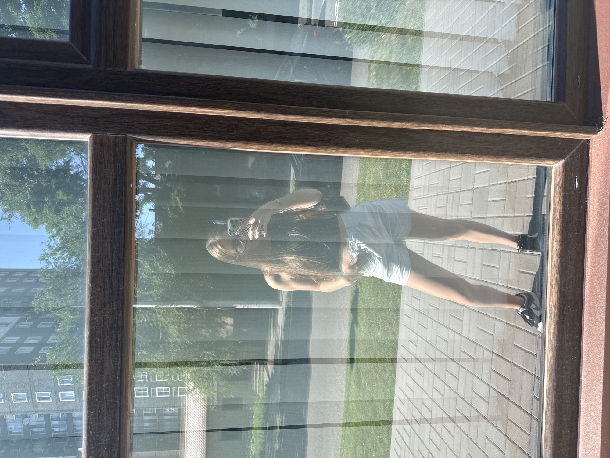 This is a vertically oriented, high-angle shot taken through a window. The window frame is dark brown and has a wood-grain texture. The glass reflects the surroundings, creating a layered image.
The primary subject is a young woman reflected in the window. She is holding a smartphone, taking a selfie. She has long blonde hair, is wearing a dark top and light-colored shorts, and black and white sneakers. Her pose is casual, with one leg slightly bent and extended. She appears relaxed.
The reflection also shows the environment outside the window. In the background, there are trees with green foliage and a bright blue sky, suggesting it is daytime. There are also glimpses of buildings with many windows, and a paved area with a brick-like pattern. A patch of green grass is visible in the lower part of the reflection.
The lighting suggests it is a sunny day, as indicated by the bright reflections and clear sky. The overall scene appears to be outdoors, possibly in a residential or urban area. There is no visible text in the image.