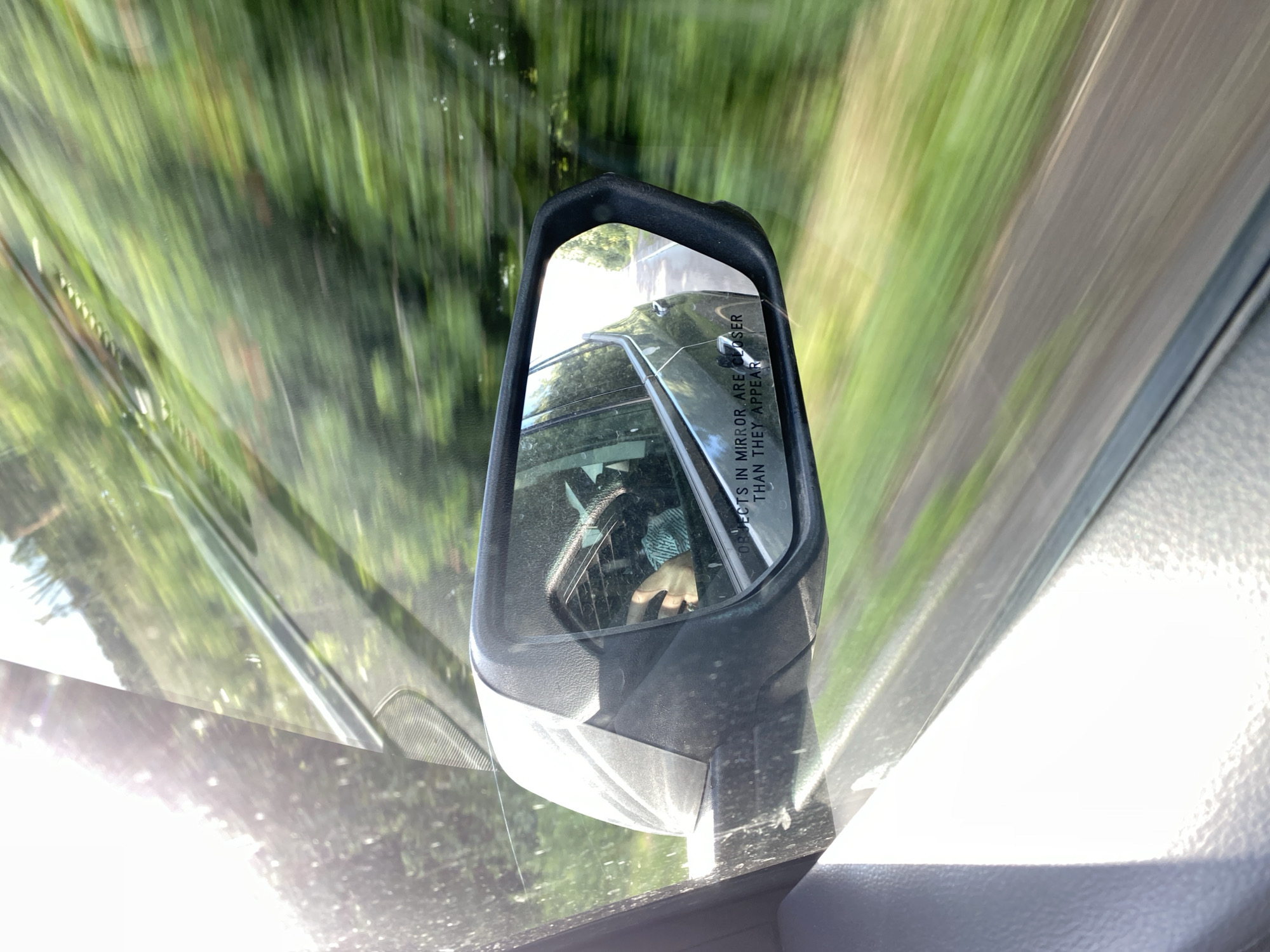 The image shows a view from inside a vehicle, looking out through the windshield and side mirror. The scene is blurred due to motion, suggesting the vehicle is in transit. Lush green foliage is visible outside, streaking across the frame, indicating a roadside environment likely with trees or bushes. The side mirror, located on the left side of the frame, reflects a portion of the car's interior and a glimpse of the outside world.
Within the reflection of the side mirror, a person's hand can be seen. The hand appears to be wearing a ring on one of its fingers. The mirror also contains text, the words "OBJECTS IN MIRROR ARE CLOSER THAN THEY APPEAR" are legible. The lighting suggests it is daytime, with sunlight creating bright areas and reflections on the windshield. The overall impression is of a drive through a natural, green landscape.