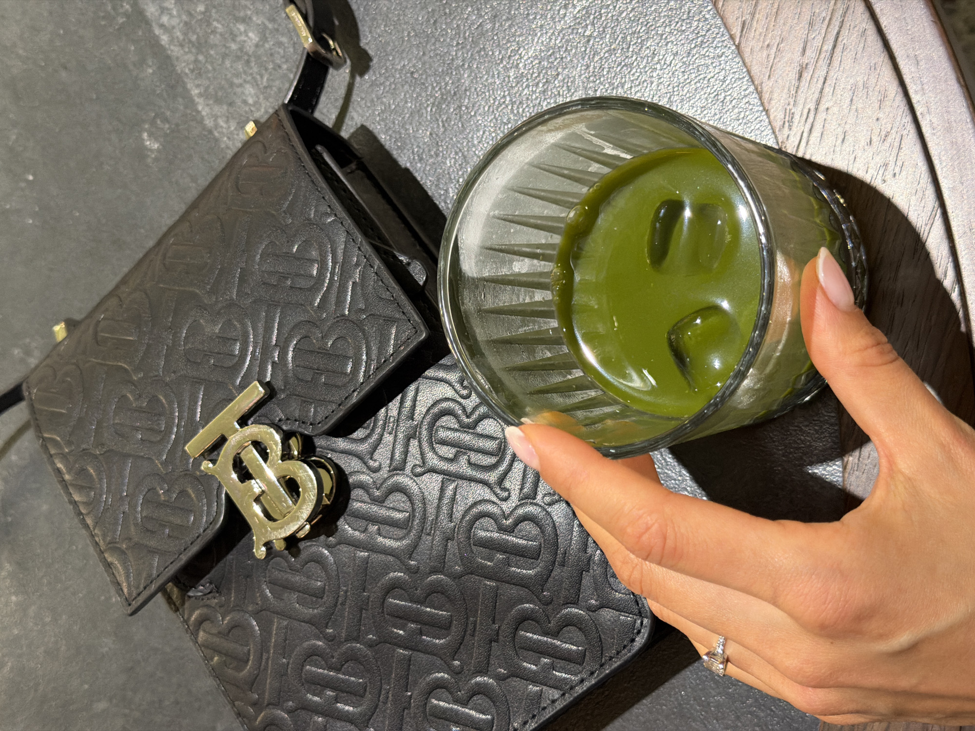 An overhead shot shows a black leather handbag with an embossed "TB" logo and a gold "TB" emblem resting on a dark textured surface. In the foreground, a glass filled with a green liquid and ice cubes is held by a person's hand. The hand has manicured nails and a ring on the index finger. The background suggests an indoor setting, possibly a cafe or restaurant in Saint Petersburg, Russia.