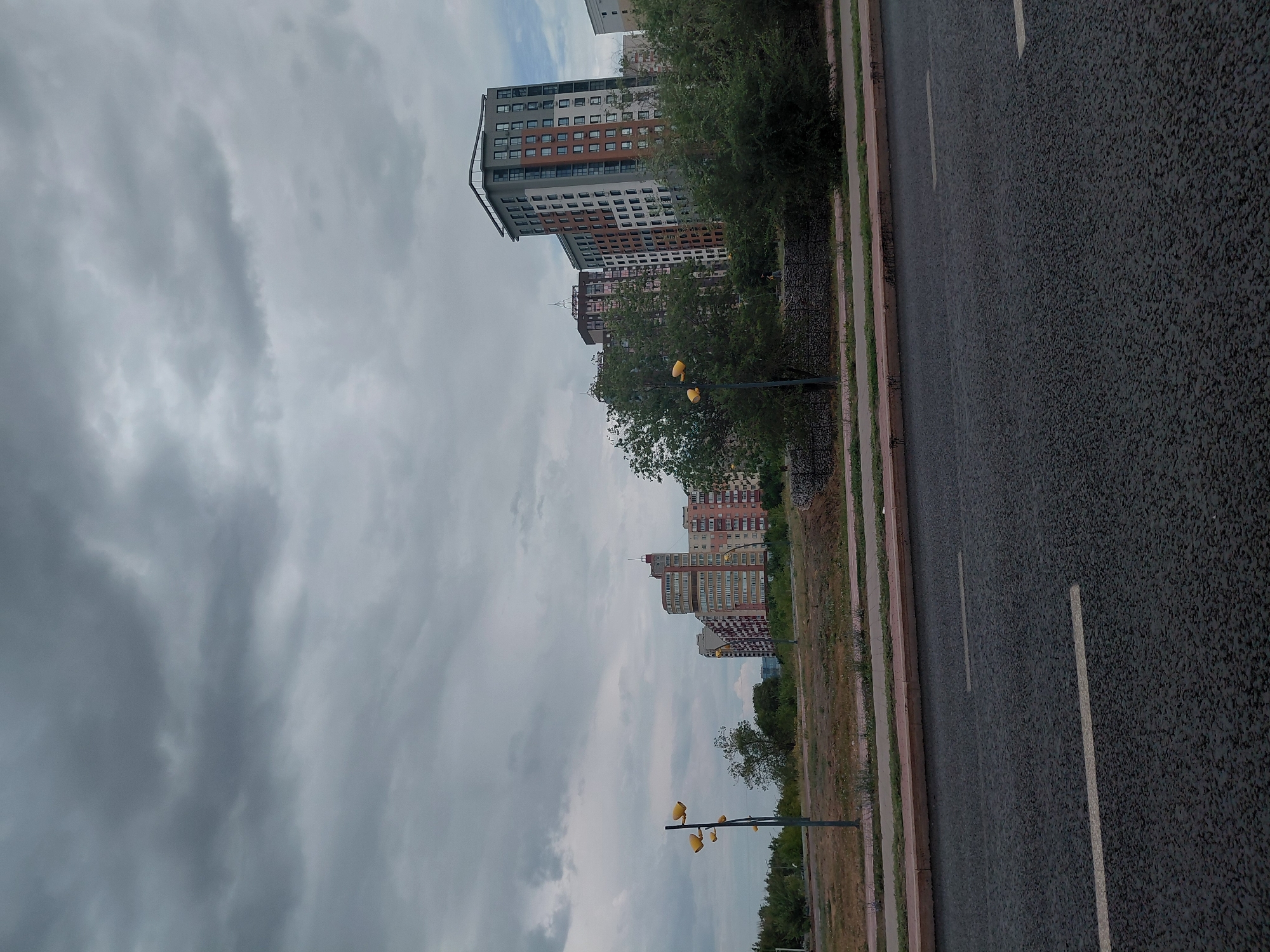 The image depicts a street scene in Qarağandı, Kazakhstan, under a cloudy sky. In the foreground, a dark asphalt road with two white lane markings is visible. A concrete curb separates the road from a dirt and grass area. Along this area, a sidewalk or pathway is present, bordered by another curb. Beyond the pathway, a row of trees and bushes lines the scene, with a low stone-filled wall acting as a barrier in places. Further back, several multi-story residential buildings of varying architectural styles and colors dominate the midground and background. One prominent building is tall and slender with a modern design, featuring many windows and a grey facade with orange accents. Other buildings exhibit more traditional designs with balconies and colorful facades. A single streetlamp with multiple yellow lights stands in the middle ground to the left. The overall impression is one of an urban residential area on an overcast day.