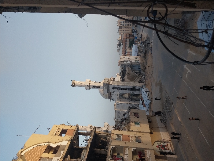 A war-torn city street displays extensive destruction. A mosque stands damaged among crumbling buildings. Debris litters the foreground. Several figures are visible on the road, some appearing to be walking and others standing near damaged structures. An overhead tangle of wires intersects the scene. The sky is a clear, pale blue.