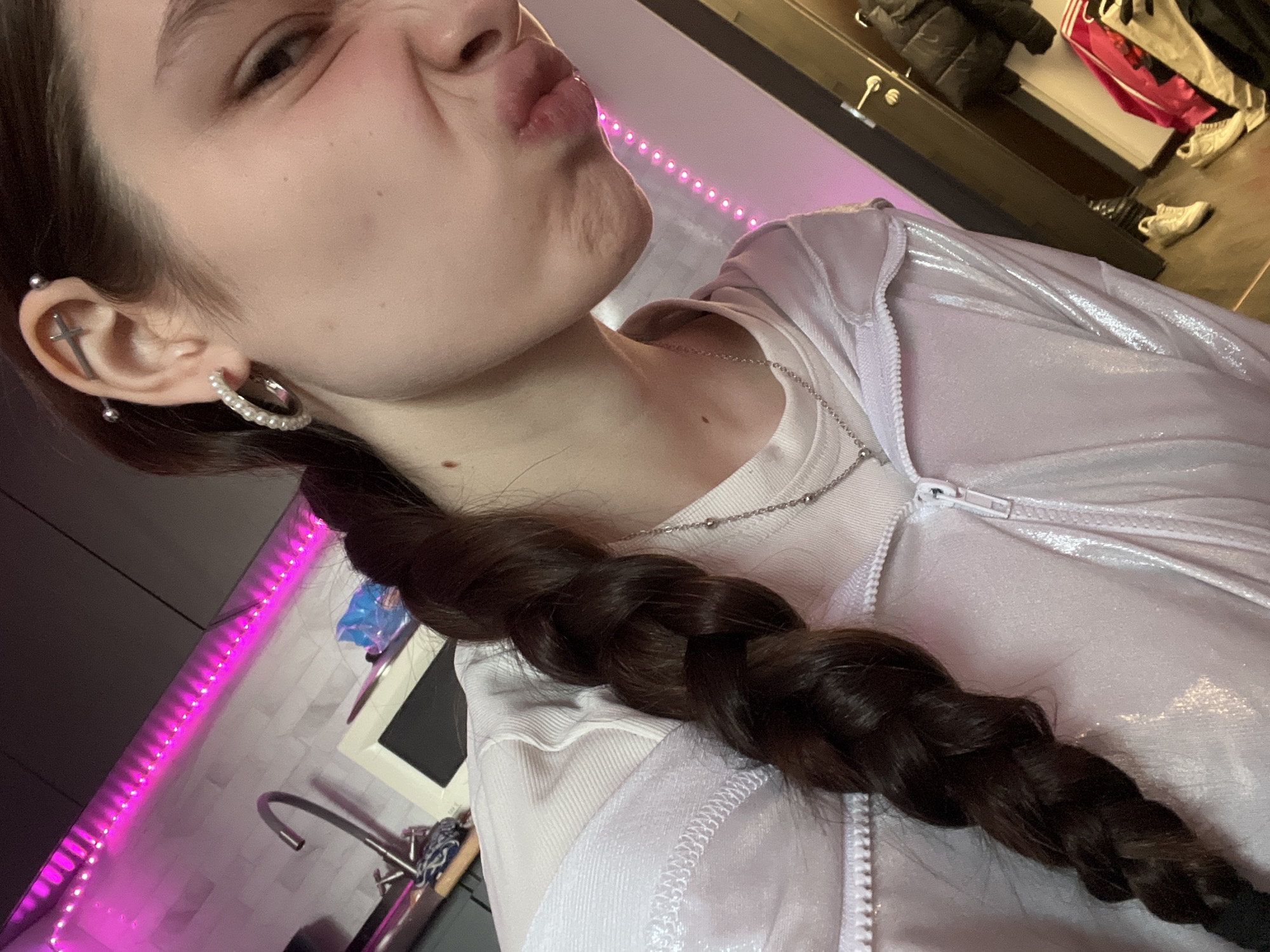 A close-up, upward-angled shot focuses on a person with a playful expression, puckered lips, and a slightly furrowed brow. The person has fair skin and dark brown hair styled in a long braid that drapes across their shoulder and chest. They are wearing a white athletic jacket with a zipper and a white T-shirt, along with a delicate silver chain necklace. The person also has an industrial piercing in their ear and a hoop earring.
The background reveals a domestic setting. To the left, part of a kitchen is visible with a sink and a decorative pink LED strip running along the edge of the counter. The area is illuminated by the pink light and overhead lighting. Behind the person, to the right, is a wardrobe with clothing items hanging inside. The location is not identifiable as Saint Petersburg, Russia, from the details provided in the image.