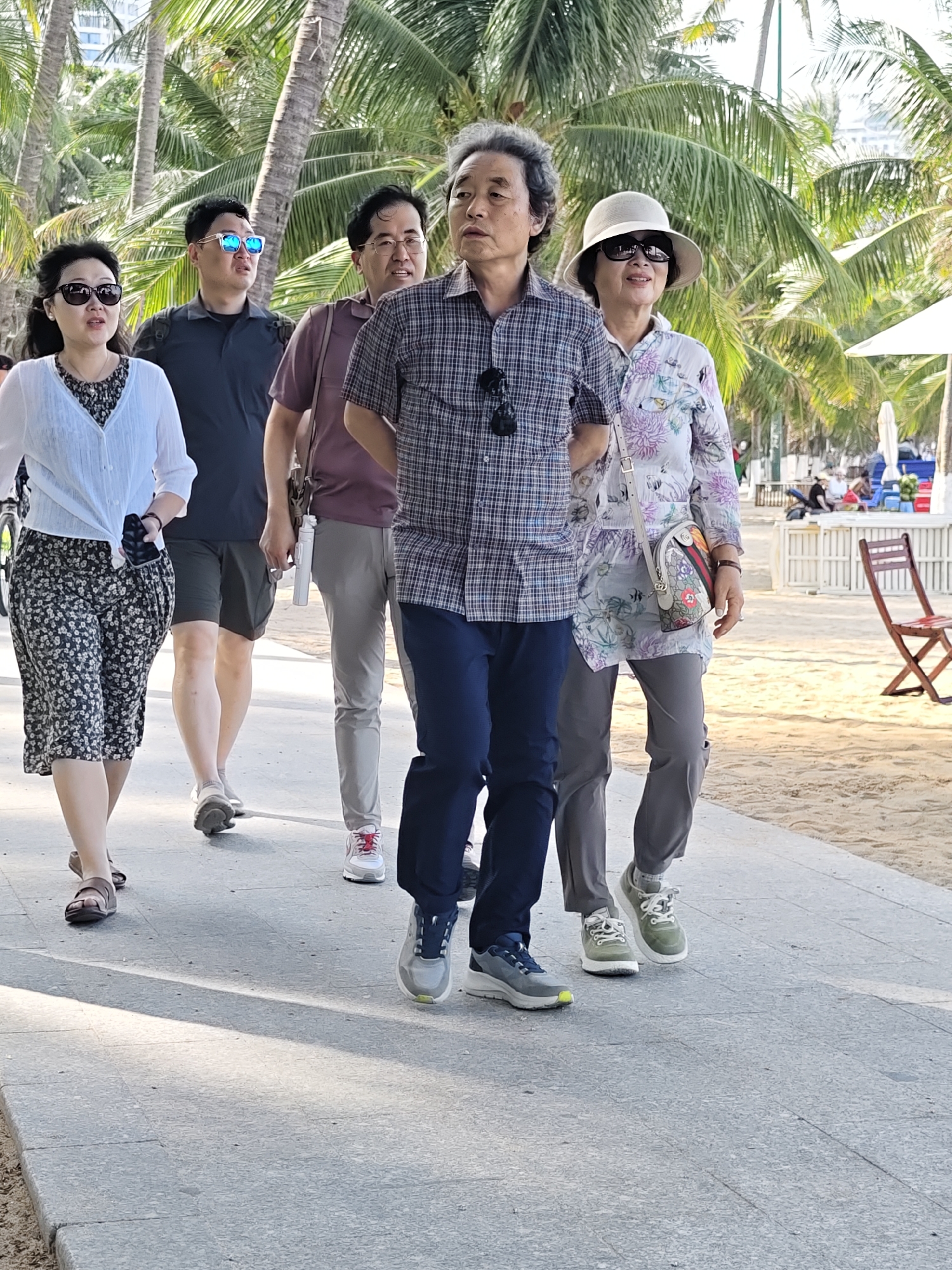 A group of five people, four adults and one woman, walk along a paved path lined with palm trees. The group consists of three men and two women. The woman in the front left wears a floral-patterned skirt and a white cardigan. To her right, a man in a dark shirt and shorts walks. Behind them, another man in a maroon shirt and gray pants walks, holding a white water bottle. Next to him, a man in a plaid shirt and blue pants is visible. The woman on the far right wears a patterned shirt and a wide-brimmed hat. The path appears to be a promenade or sidewalk, with a sandy beach visible to the right, and palm trees and buildings in the background, suggesting a location in a coastal city like Nha Trang Ward, Vietnam. The scene captures the group in mid-stride, appearing to be on a leisurely walk.