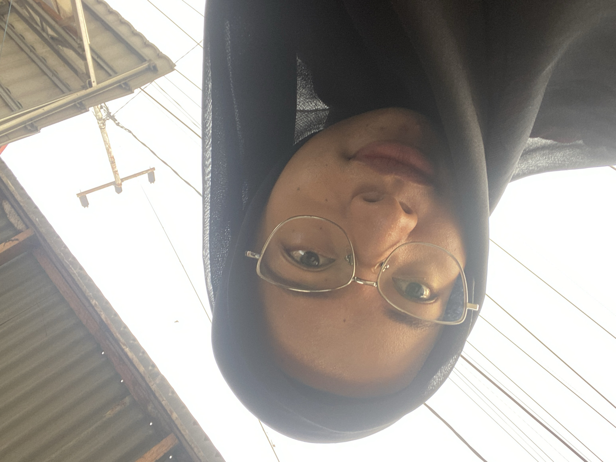 A person wearing glasses and a hijab is looking at the camera. The image is taken from a low angle, looking up at the person and the sky. There are power lines and a corrugated metal roof in the background. The scene appears to be outdoors in an urban or semi-urban environment. The location is identified as Air Molek, Indonesia.