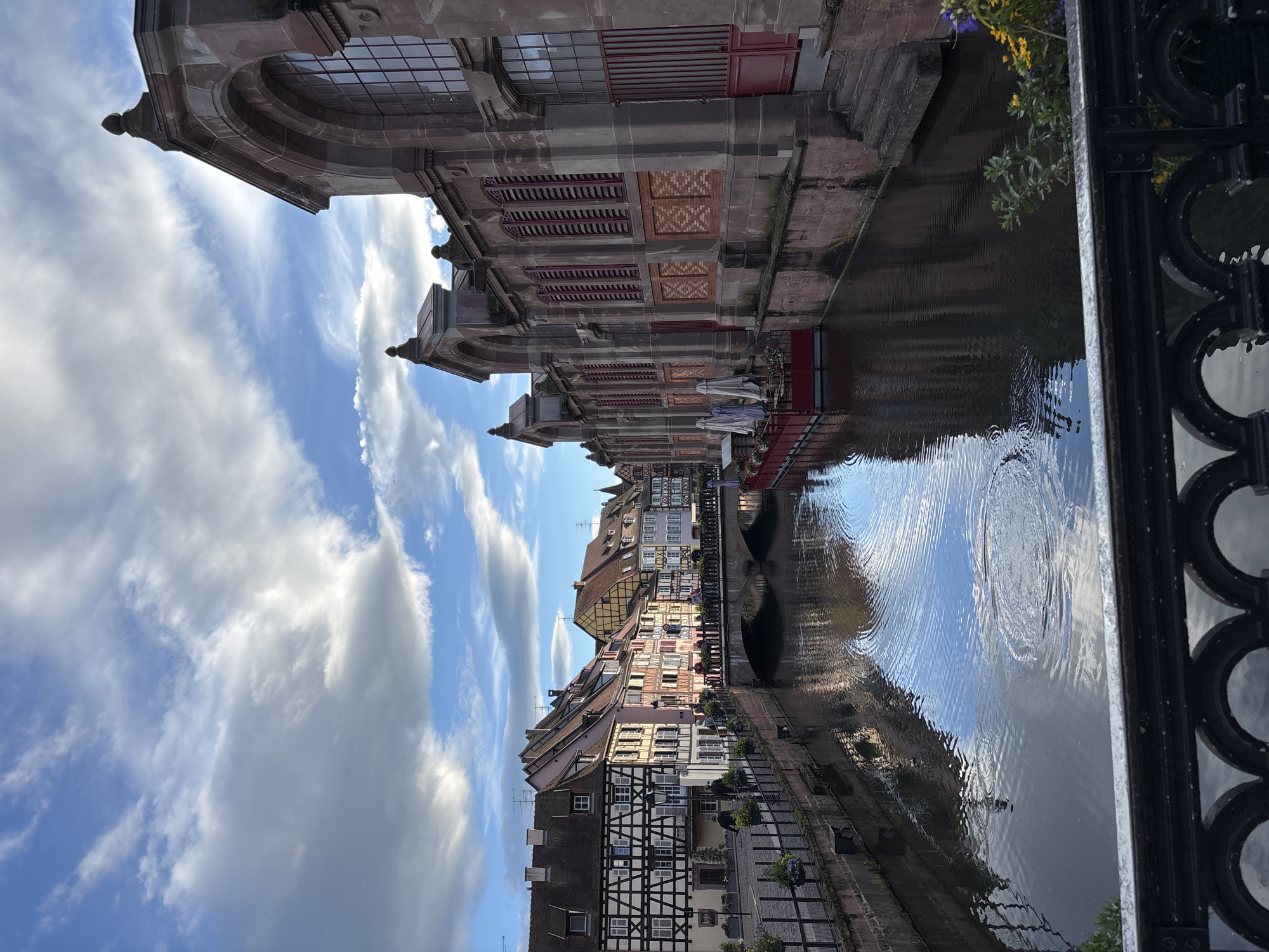 The image depicts the Lauch river in Colmar, France, a picturesque canal lined with historic buildings. On the left side, a row of half-timbered houses with colorful facades and steep roofs are visible, characteristic of the Alsatian architectural style. A stone bridge spans the canal, with people walking across it. On the right side of the canal, a large, imposing stone building with arched windows and red shutters stands prominently. Several closed white umbrellas are set up in front of this building, suggesting a cafe or restaurant. The canal itself is dark and reflective, mirroring the sky and the surrounding architecture. The sky above is partly cloudy, with patches of blue sky visible between dramatic white clouds. A black metal railing runs along the bottom of the foreground, partially obscuring the view of the canal and the buildings. The overall scene conveys a sense of charming European urban landscape bathed in natural light.