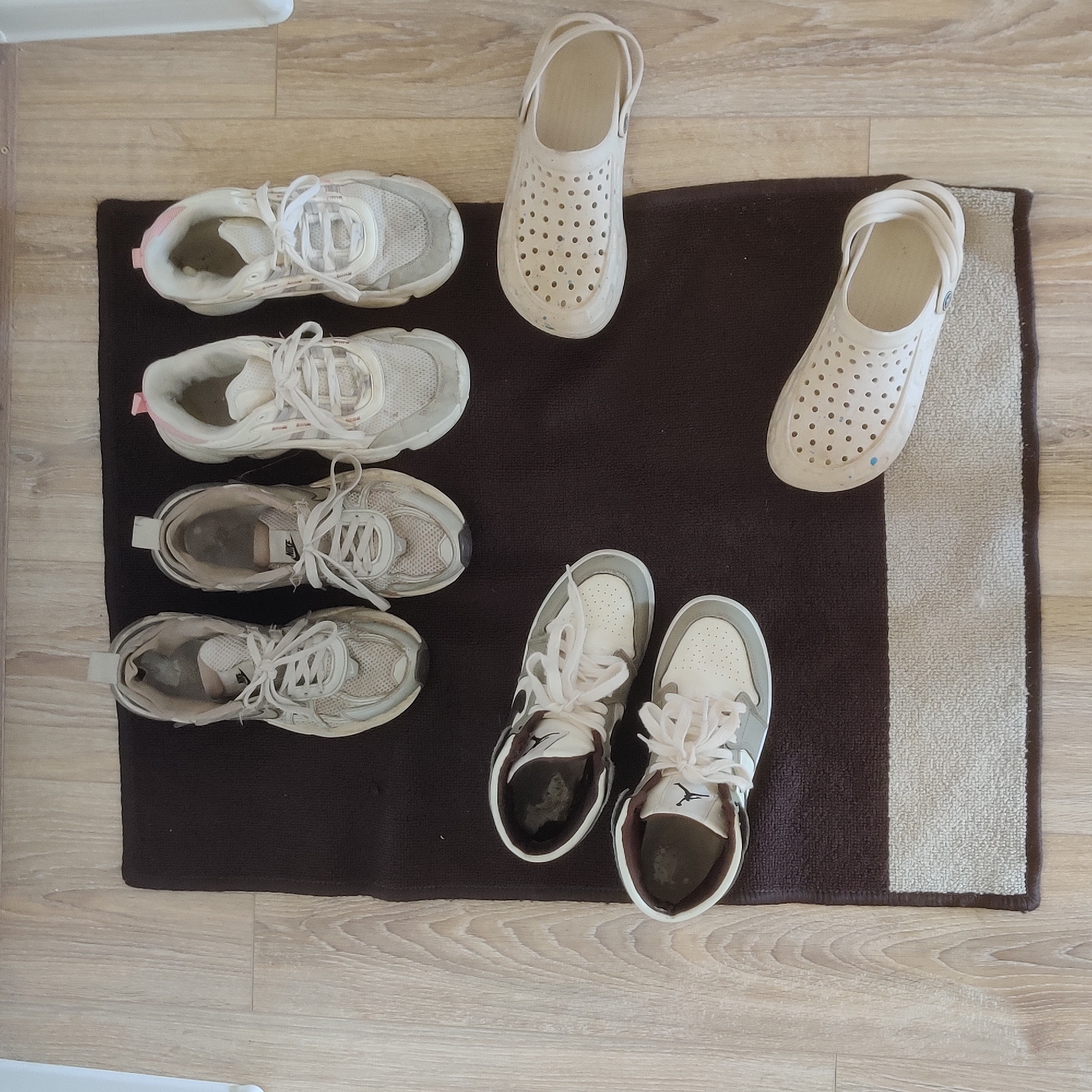 The image shows several pairs of shoes and a doormat on a wooden floor. In the top half of the image, there are three pairs of athletic shoes, two white and one with pink accents, arranged in a row. Below these, on the left side of the doormat, are two more pairs of athletic shoes, both white with brown accents. To the right of the doormat, there are two white perforated slide-style shoes. The doormat itself is dark brown with a lighter beige stripe along the bottom edge. The wooden floor has a light brown, wood-grain pattern. The scene appears to be indoors, likely a home, and there is no information to suggest a location in Yekaterinburg, Russia.