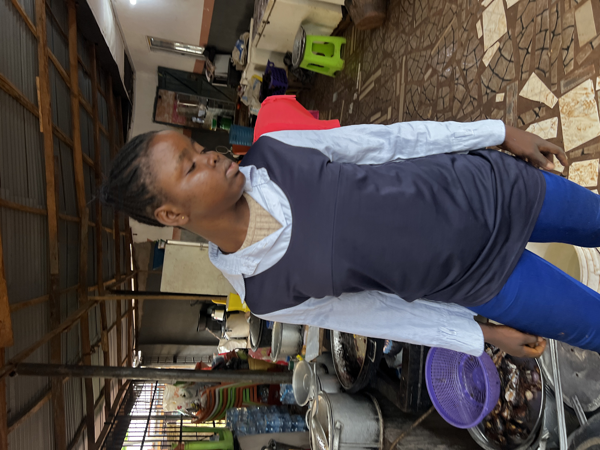 The image depicts an outdoor or semi-outdoor market environment in Nkpor, Nigeria. In the foreground, a young woman with dark skin and braided hair stands facing to the right, looking down. She is wearing a light blue pinstripe shirt with a dark sleeveless top over it, and bright blue trousers. Her left hand is visible near a purple colander filled with food items.
The background reveals a busy marketplace setting. Various cooking utensils and food preparation equipment are scattered around, including large metal pots, a wok-like pan with cooking food, and buckets. Several plastic crates and bottles of water are stacked in the background. The structure of the market is rudimentary, with a corrugated metal roof supported by wooden beams and poles. The floor is tiled with a mosaic pattern. The overall impression is of a functional, working market area.