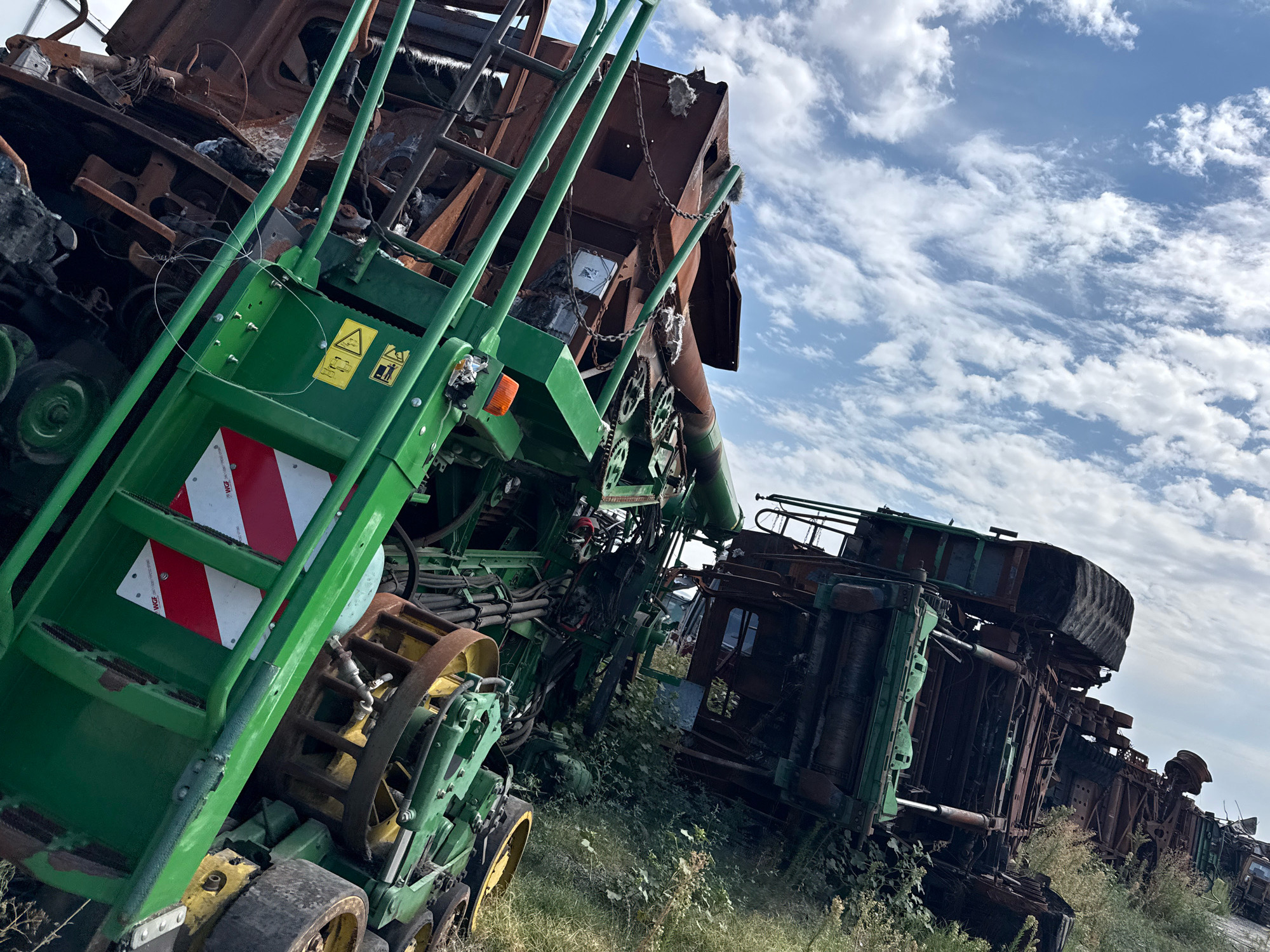 In Veselinovo, Bulgaria, a scene of destruction unfolds with several large agricultural machines, seemingly harvesters, heavily damaged and overturned. The machinery, primarily green and rusted brown, is entangled with grass and weeds, suggesting abandonment or a recent catastrophic event. One harvester in the foreground is particularly visible, its green frame and components sharply contrasted against the weathered metal. Red and white reflective markings are present on some parts of the machinery. The sky above is filled with a mix of white, puffy clouds and lighter wisps against a bright blue backdrop. The overall impression is one of disarray and ruin within an outdoor, natural environment.