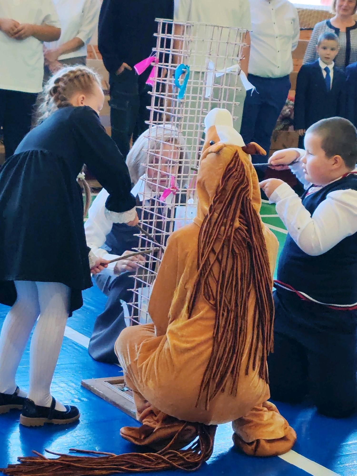 The image depicts an indoor scene, likely a gymnasium or school hall, with a blue floor marked with white lines. In the foreground, a child in a brown horse costume with long brown fringe hair is crouching. To the right, another child, a boy in a dark vest over a white shirt and tie, is standing and appears to be interacting with the costume. To the left, a girl in a black dress and white tights is also partially visible, with her back to the camera. Behind them, several adults and children are standing, some dressed in school uniforms. A white mesh structure with pink and blue ribbons is positioned centrally in the image. The overall impression is of a school event or performance taking place in a public building within the city of Chukhlom, Russia.