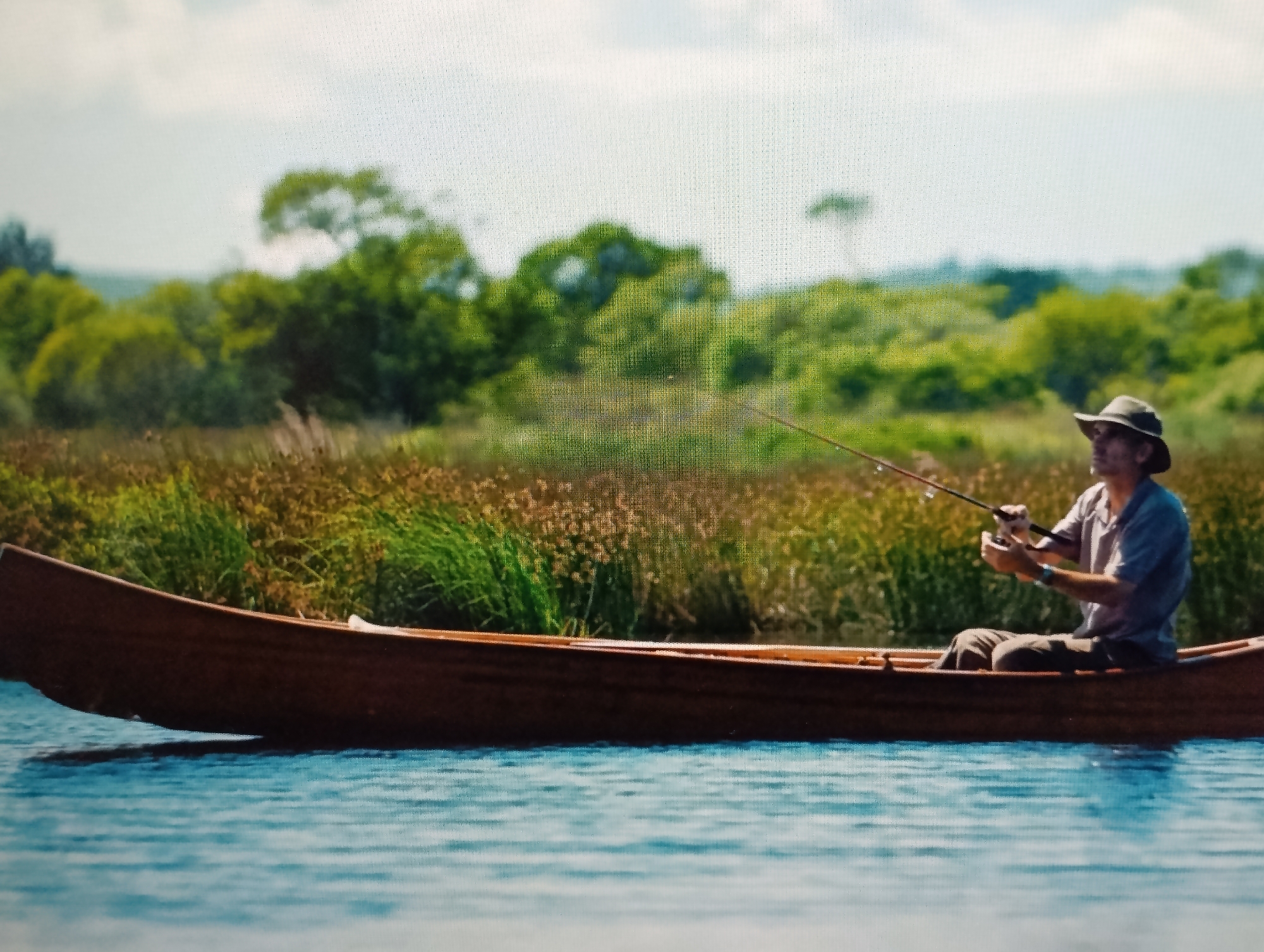 A man wearing a hat and a blue shirt is fishing from a wooden canoe on a body of water. He is holding a fishing rod and appears to be reeling in his line. The canoe is brown and is partially submerged in the blue water, casting a shadow. The background features lush green trees and tall brown grass, suggesting a natural, possibly rural environment. The sky is bright and partly cloudy. The image appears to be taken on a sunny day.