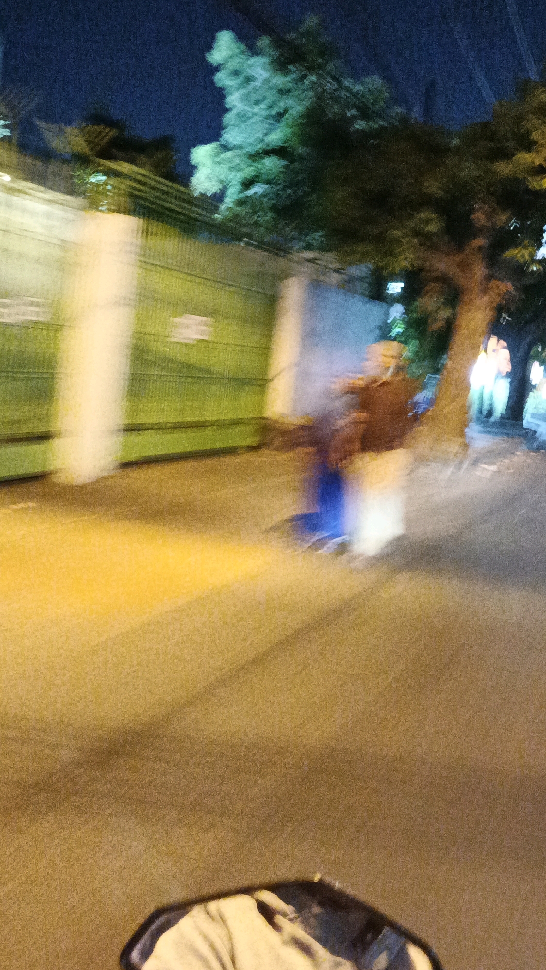 The image captures a blurred nighttime street scene, likely in Surakarta, Indonesia, given the context. A person is visible on a vehicle, appearing to be in motion, with their form indistinct due to the blur. They are wearing a dark upper garment and have what looks like a blue object behind them. The background features a long, green fence with vertical white elements, suggesting it might be part of a property boundary or a building. Trees with foliage, illuminated by artificial light, are also present, casting shadows on the road. The road surface is visible, reflecting yellow light from street lamps, and a portion of a vehicle's mirror or handlebars is in the foreground, further indicating movement. The overall impression is one of a busy urban environment at night, with vehicles and pedestrians passing by.