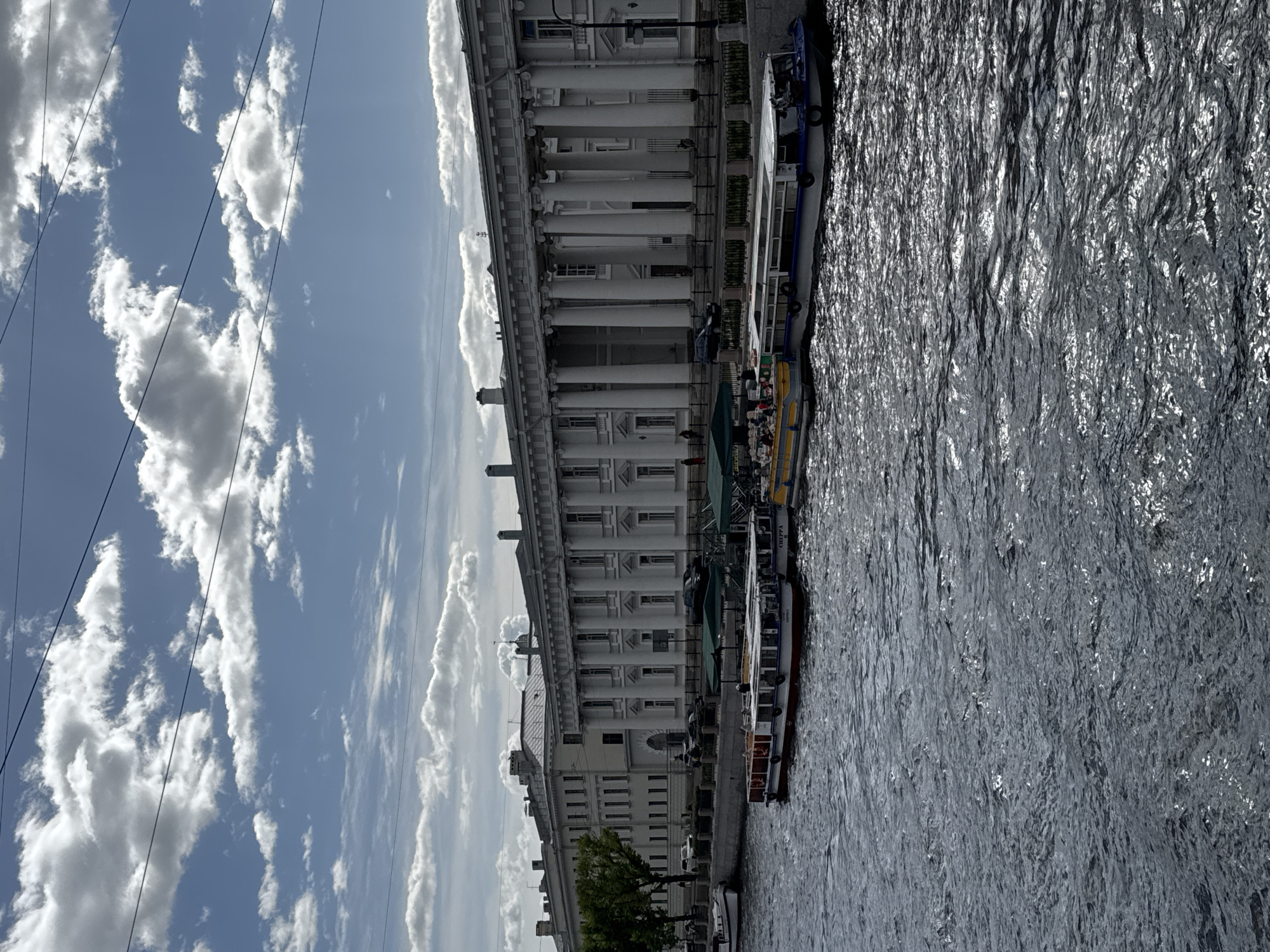 The image shows a waterfront scene in Saint Petersburg, Russia, dominated by a classical building with a long colonnade, likely a palace or government structure, bordering a canal or river. Several boats are moored along the embankment, presumably tour boats. The water is rippled, reflecting the light. The sky is blue with scattered white clouds. Power lines are visible against the sky.