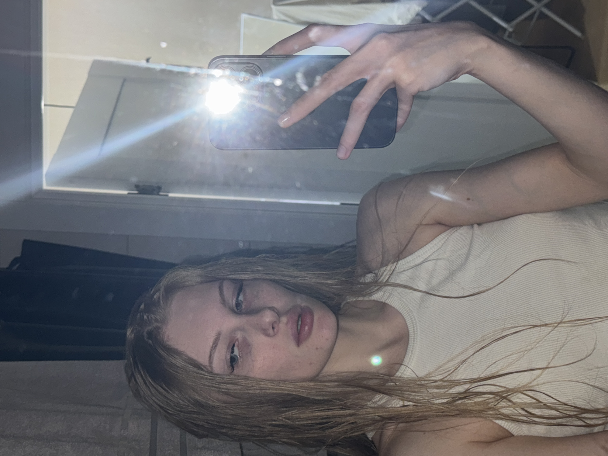 A young fair-skinned person with long blonde hair wearing a cream tank top takes a selfie with a phone. The environment is indoors, possibly a bedroom, with a window and a dark object partially visible in the background. There are no indicators to define the location as Perm, Russia.