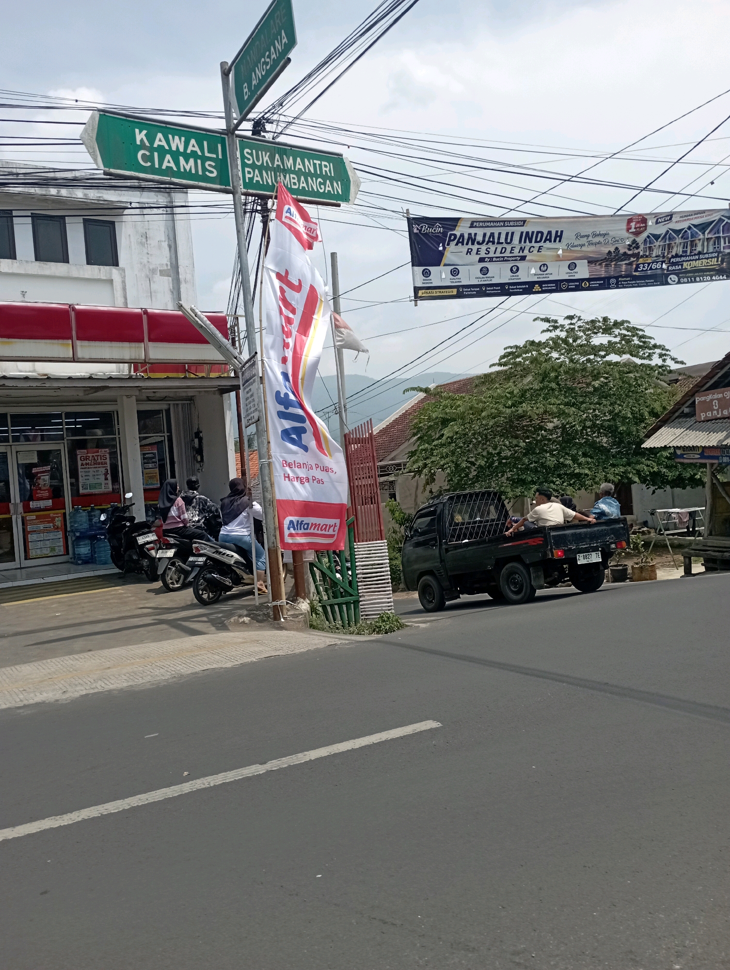 The image shows a street scene in Panjalu, Indonesia, with an Alfamart convenience store on the left. Several motorcycles are parked in front of the store, and a few people are standing near them. A white flag with the Alfamart logo is prominently displayed. A banner advertising "Panjalu Indah Residence" hangs overhead, along with numerous electrical wires. In the background, there are houses and trees, and a small black pickup truck is driving away from the camera, carrying passengers in its bed. Street signs indicate directions to "Kawali Ciamis" and "Sukamantri Panjumbangan".