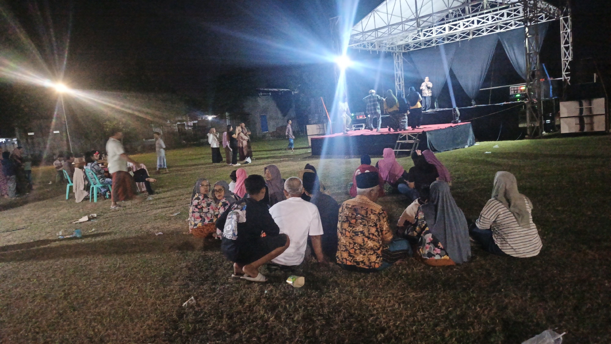 A crowd of people are gathered on a grassy field at night, watching a performance on a stage. The stage is lit by bright lights, and there are speakers set up on either side. The audience is seated on the ground, with some people standing in the background. The scene appears to be an outdoor event, possibly a concert or festival, in Ketemasdungus, Indonesia. The people are dressed in casual clothing, and many of them are wearing headscarves. The overall atmosphere is one of community and shared experience.