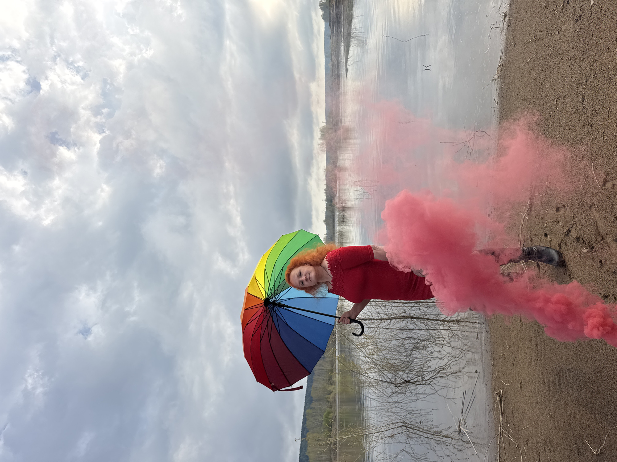 A woman stands at a lake edge on sandy ground, holding a rainbow umbrella in her left hand and a pink smoke grenade in her right. She wears a red dress and black boots. The lake water is visible behind her, with bare tree branches emerging from the water's edge. In the background, there are distant trees. The sky is cloudy. The location is Noviy, Russia.