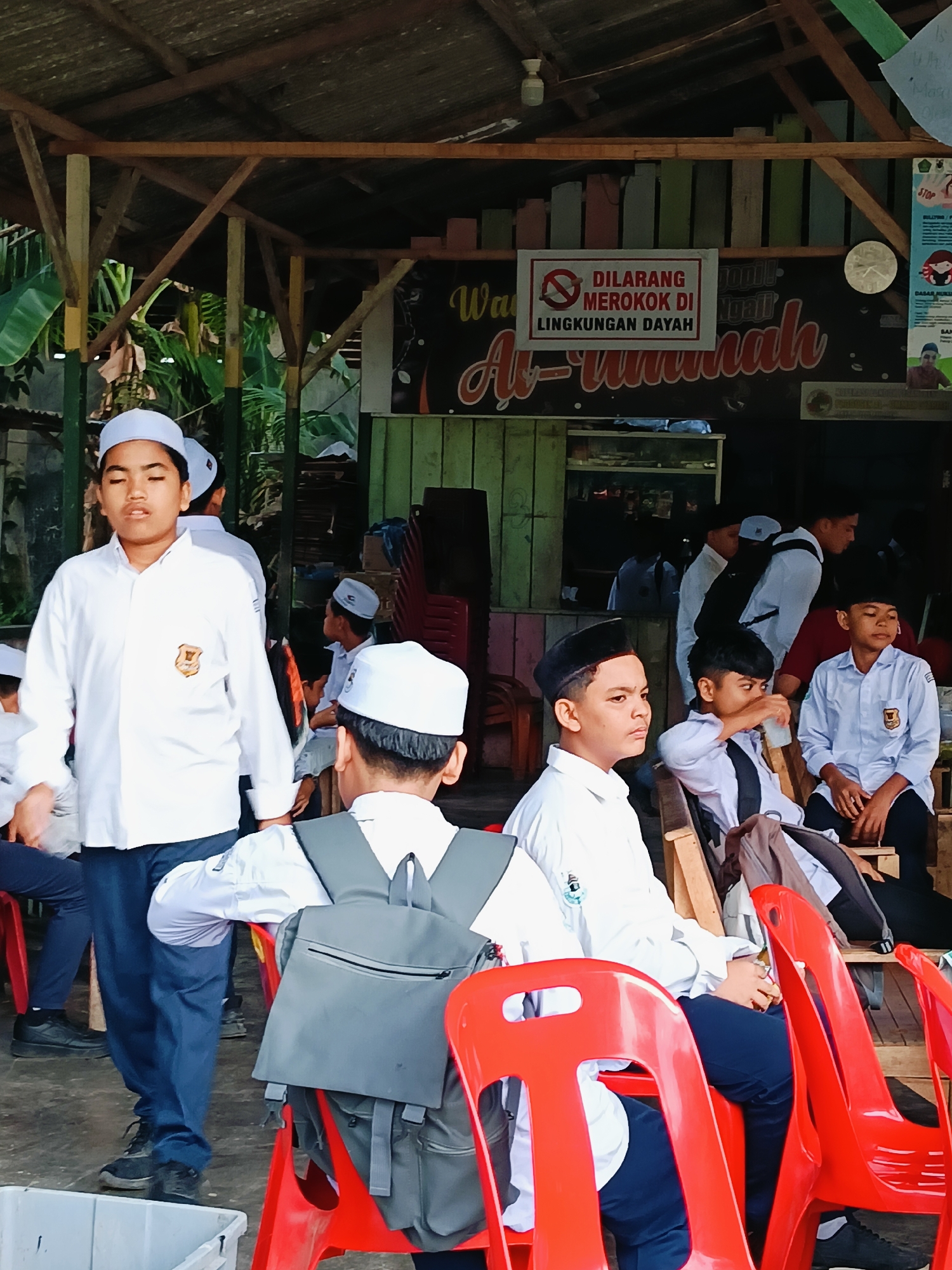 The image shows a group of adolescent males, possibly students, in Lhokseumawe, Indonesia. Most are seated on red plastic chairs under a covered structure. All are wearing white shirts with blue pants or shorts and white or black caps. A sign reading "DILARANG MEROKOK DI LINGKUNGAN DAYAH Al-Ummah" (NO SMOKING in the Al-Ummah Dayah environment) is visible in the background. The surrounding area appears to be semi-outdoor, with greenery and other buildings visible.