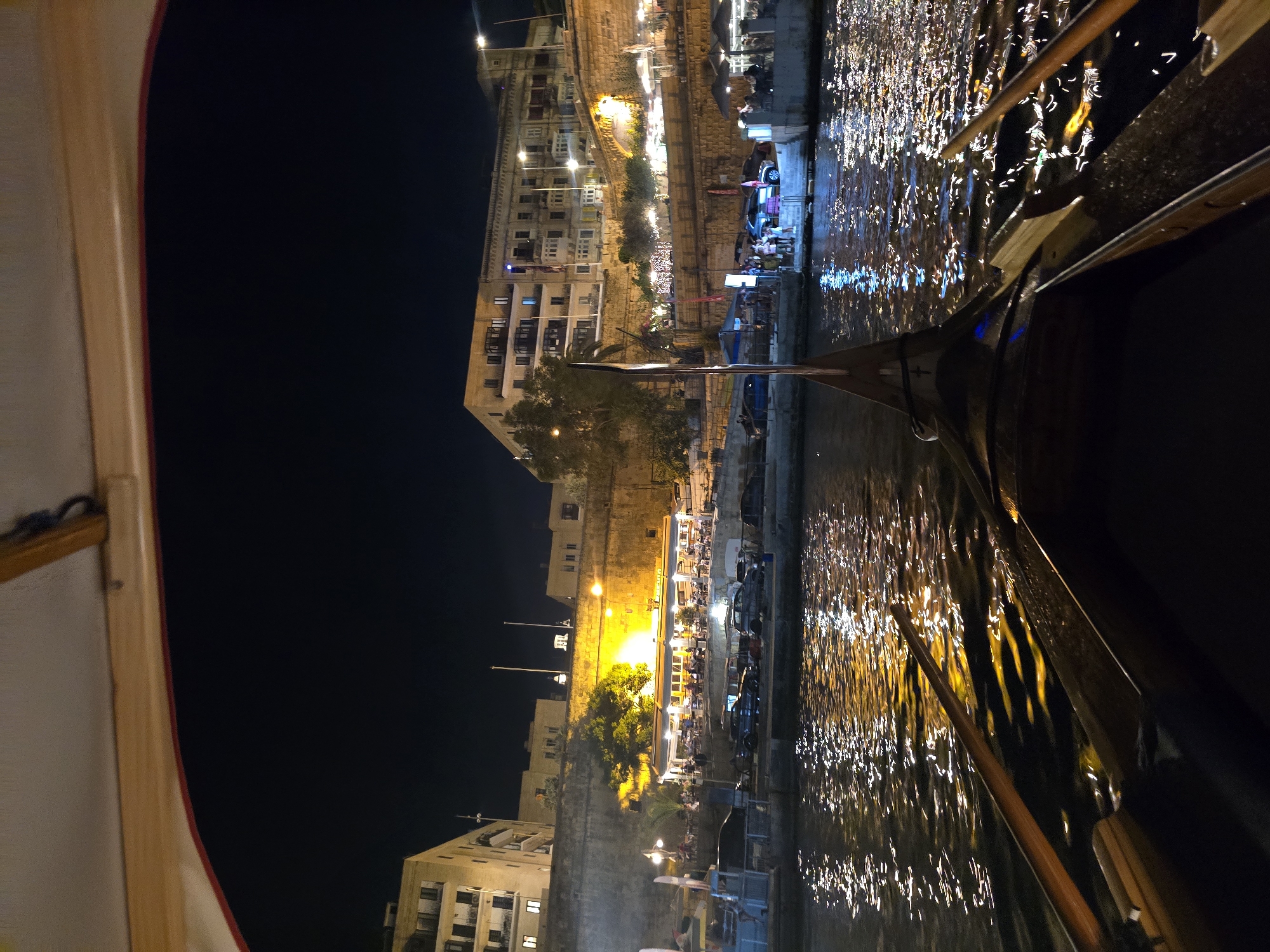 This image captures a nighttime view from a gondola on the water in Valletta, Malta. The perspective is from inside the gondola, looking forward. The water is dark with shimmering reflections of lights from the city. In the background, the illuminated city walls of Valletta are visible, with buildings and restaurants lining the waterfront. Several people are seated at tables outside a restaurant under bright lights. Cars are parked along the embankment. The foreground shows the wooden structure of the gondola and two oars. The sky is dark, indicating it is nighttime.