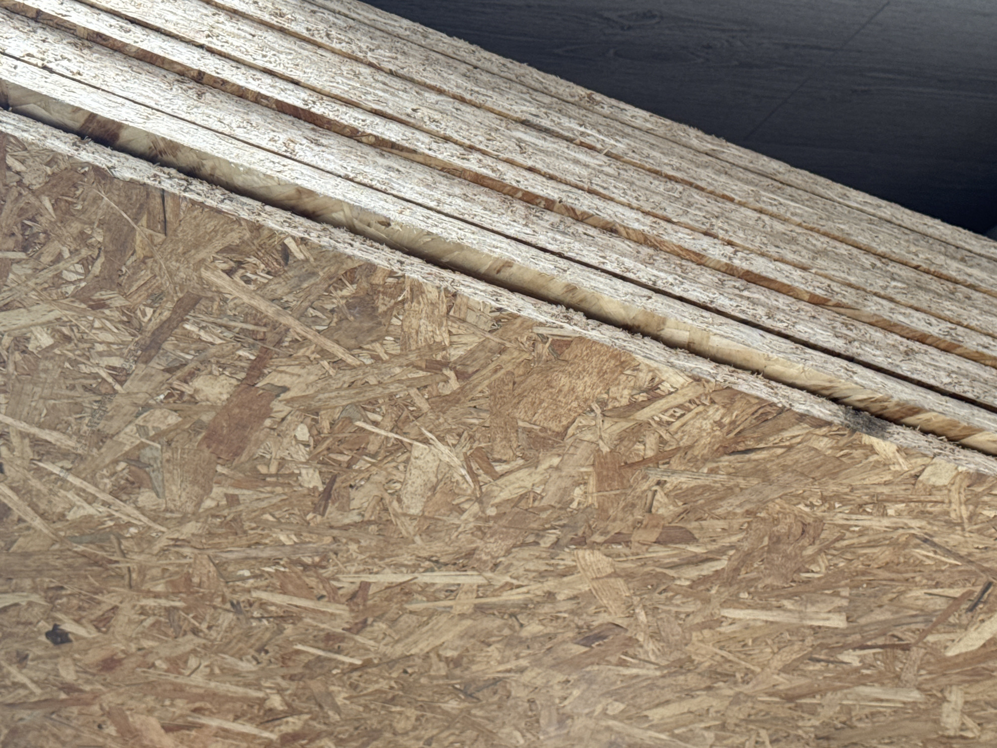 The image shows a close-up view of oriented strand board (OSB) panels stacked together. The OSB has a rough texture composed of wood chips and fibers compressed and bonded with resin. To the right of the OSB, a stack of long, thin, light-colored wooden planks are visible, also positioned diagonally. The background is a dark, textured surface, possibly a wall or floor. The scene appears to be an indoor storage area, perhaps a construction site or a workshop, given the materials. The location is specified as Яромiнскi сельскi Савет, Belarus. No people or actions are depicted.